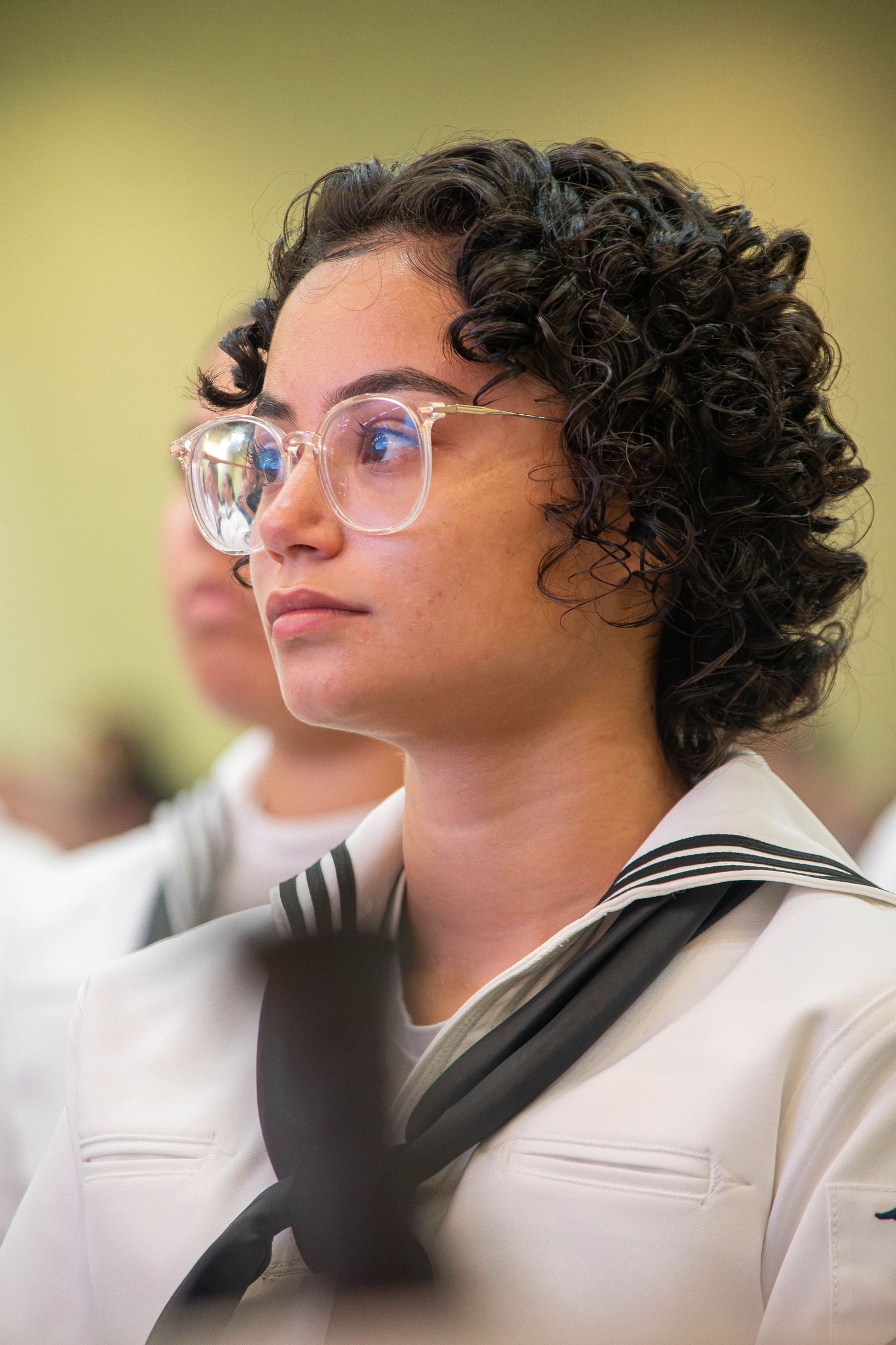 Female Navy sailor with curly hair and glasses in white uniform at military ceremony.