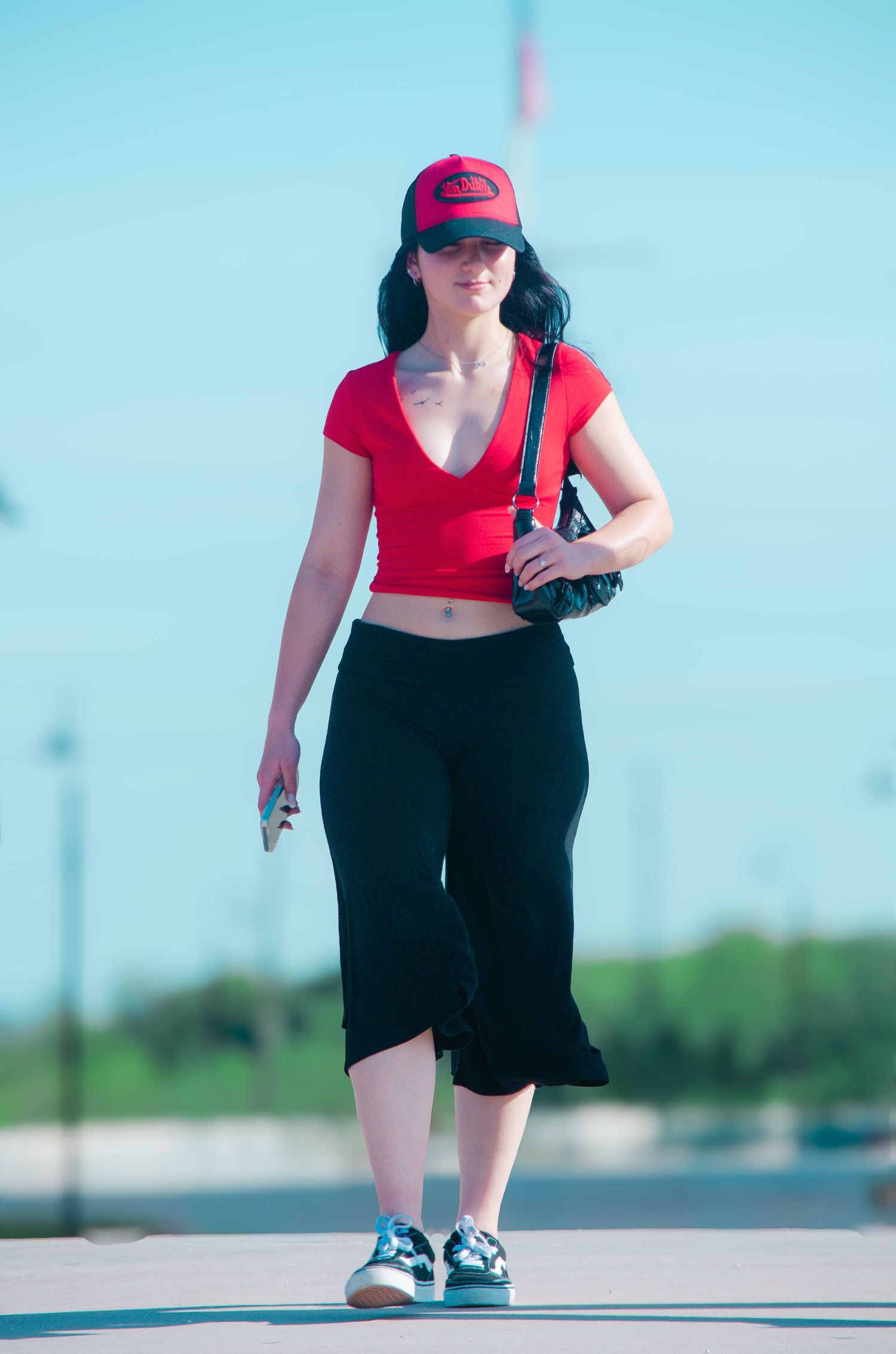 Young woman in red crop top, black capris, and red cap walking outdoors on a sunny day.