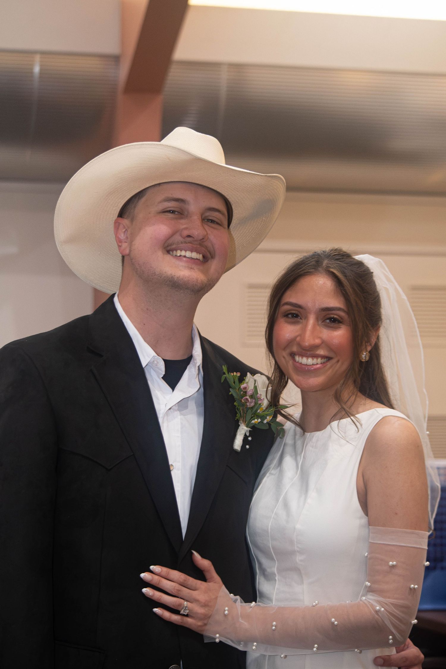 Smiling bride and groom in white cowboy hat posing together at wedding ceremony indoors.