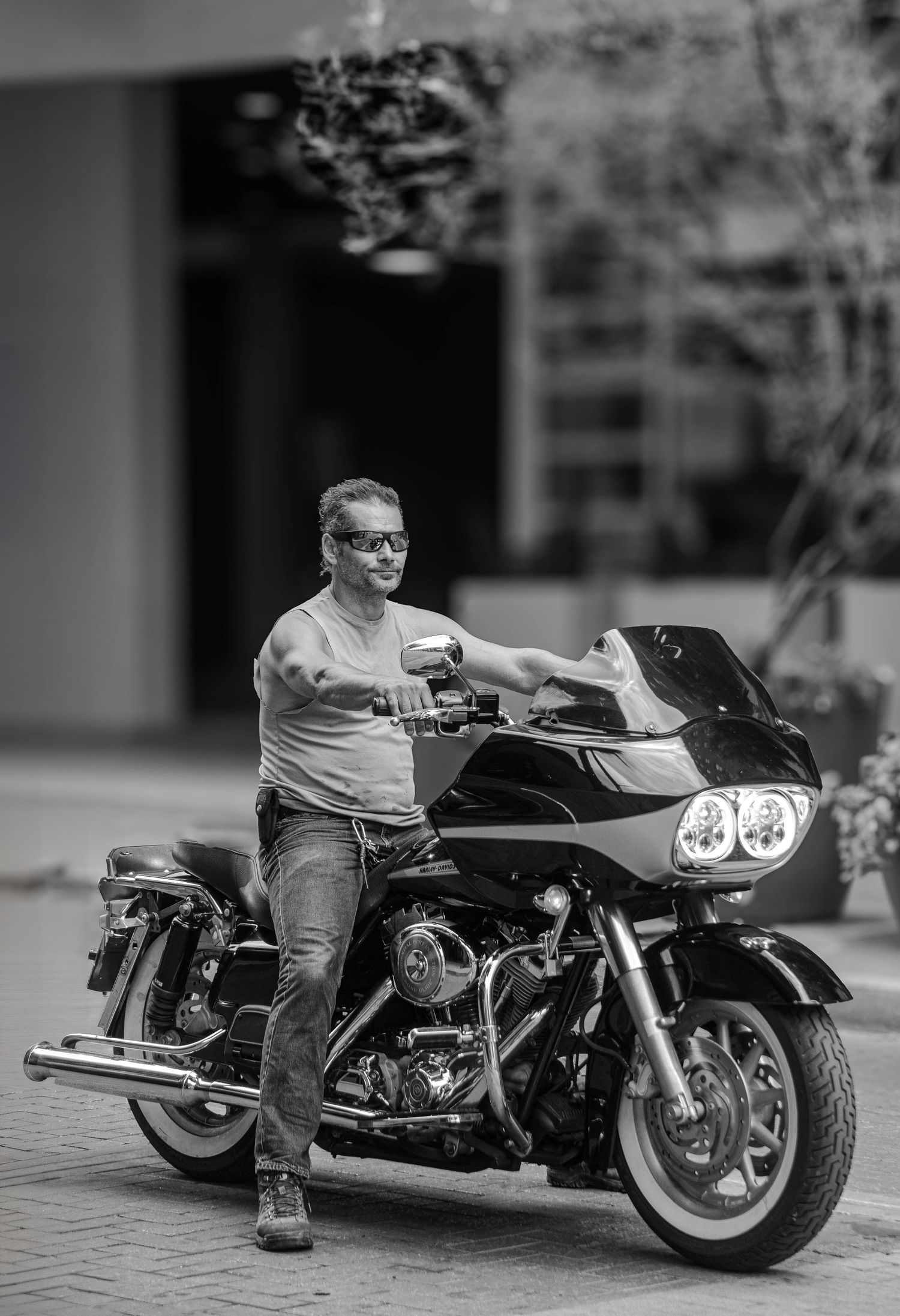 Black and white portrait of a man in sunglasses leaning on a Harley-Davidson motorcycle.