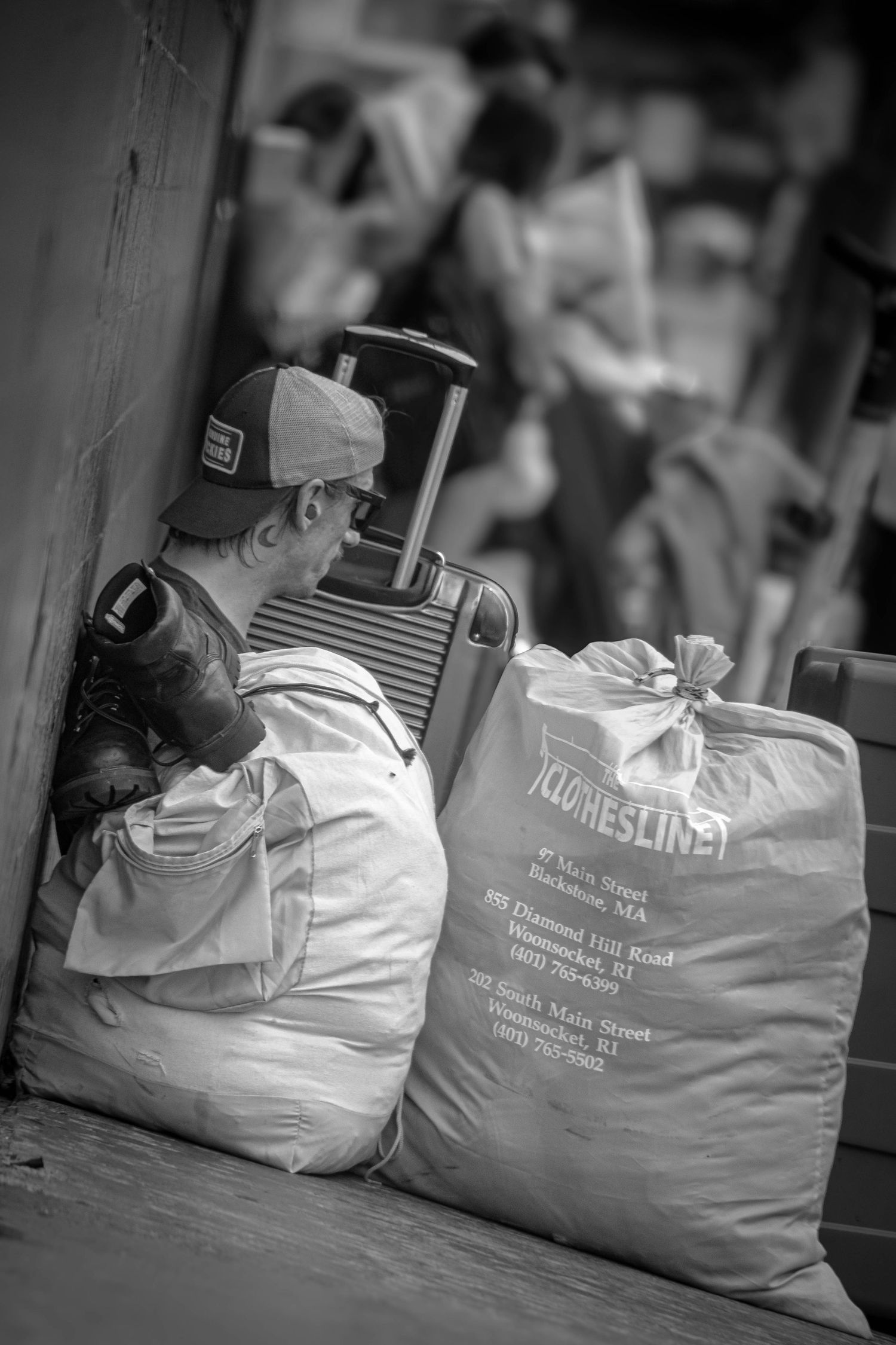 Black and white photo of a man sleeping against a wall beside a suitcase and laundry bags.