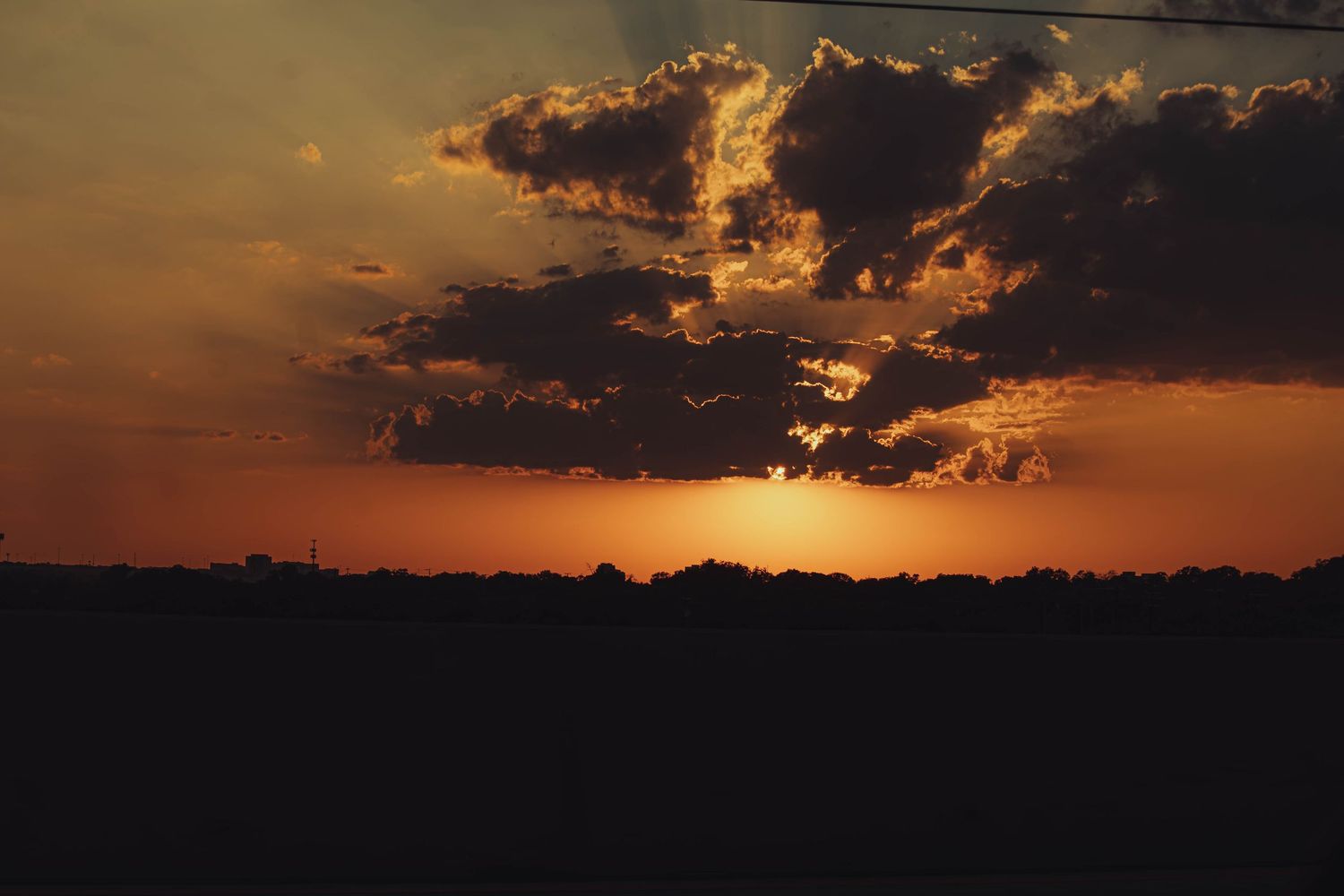 Dramatic orange sunset with dark clouds silhouetting a flat treeline on the horizon.