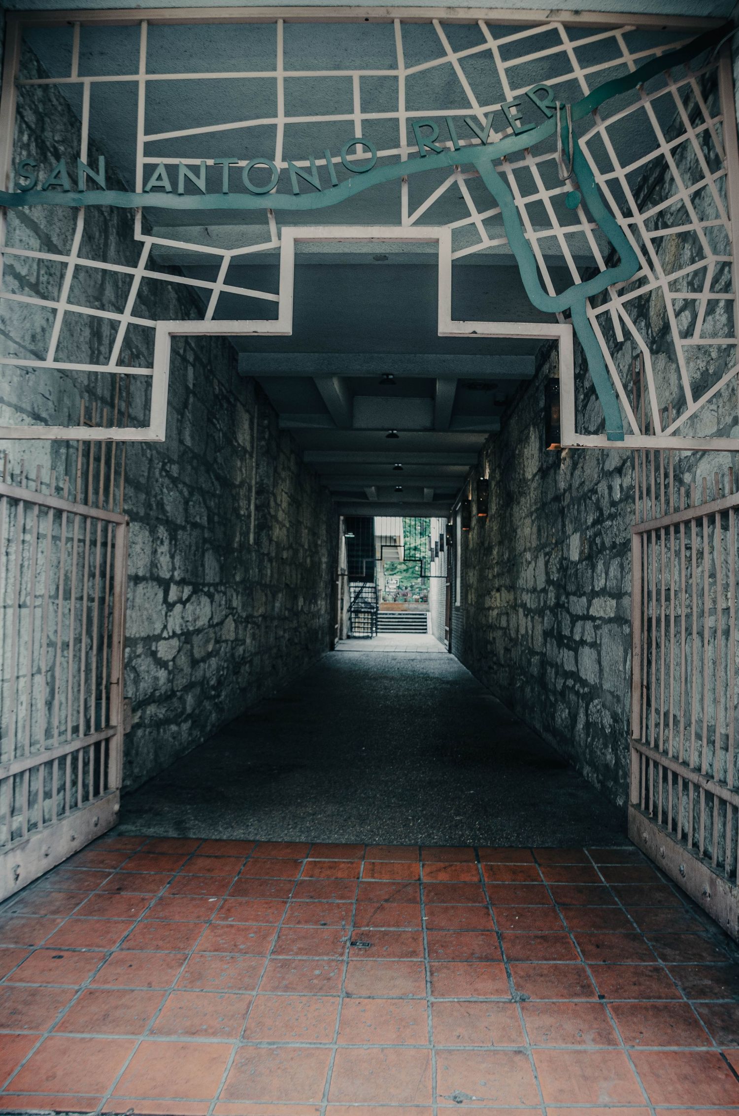 Narrow stone tunnel passageway with San Antonio River map mural and iron gates.