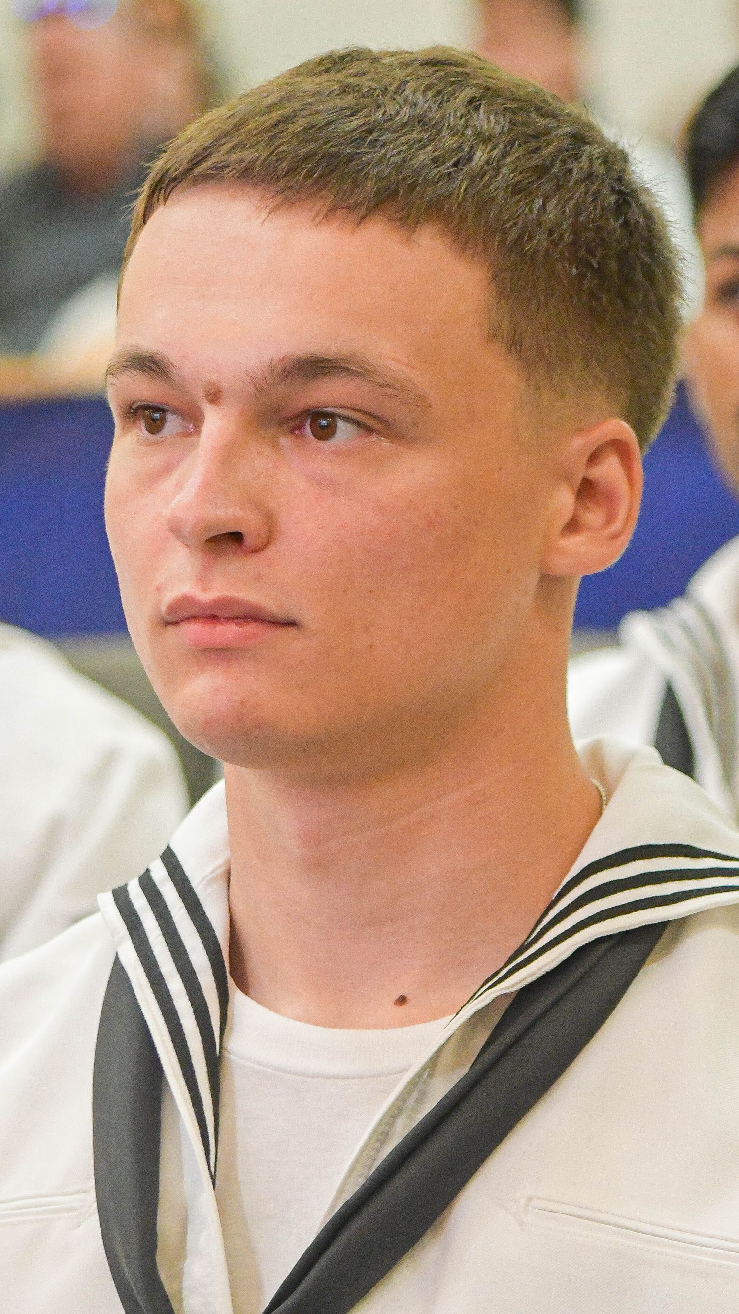 Young male Navy recruit in white sailor uniform attends military training graduation ceremony indoors.