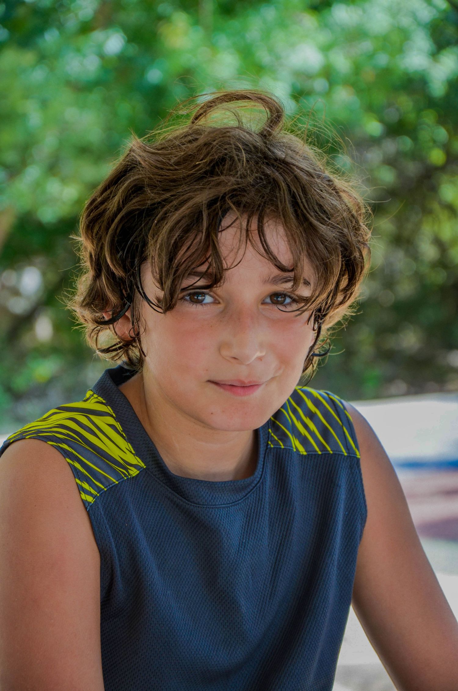 A young boy with tousled brown hair and blue eyes wearing a dark sleeveless shirt outdoors.