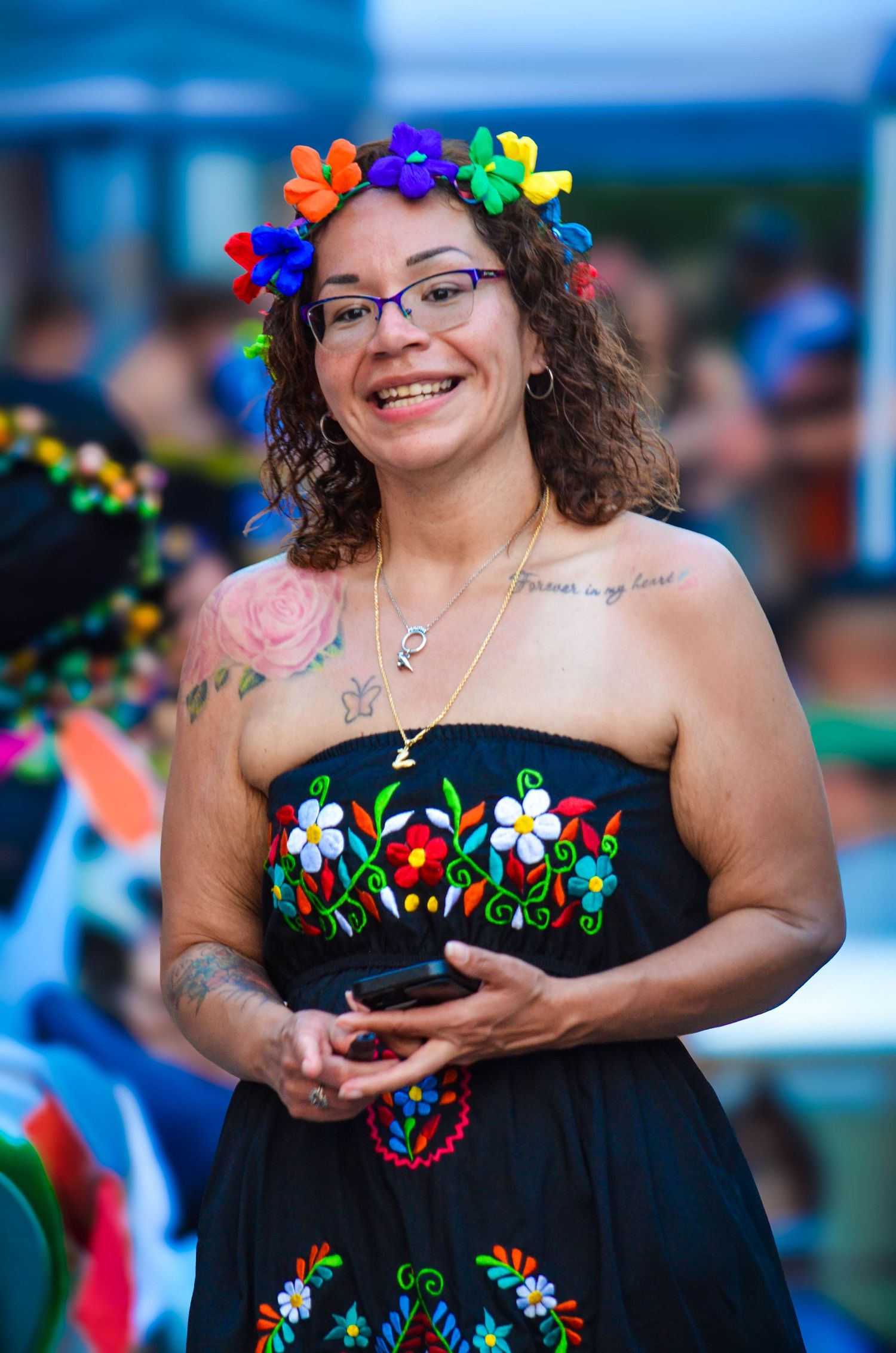 Smiling woman in floral embroidered strapless dress wearing a colorful flower crown at an outdoor festival.