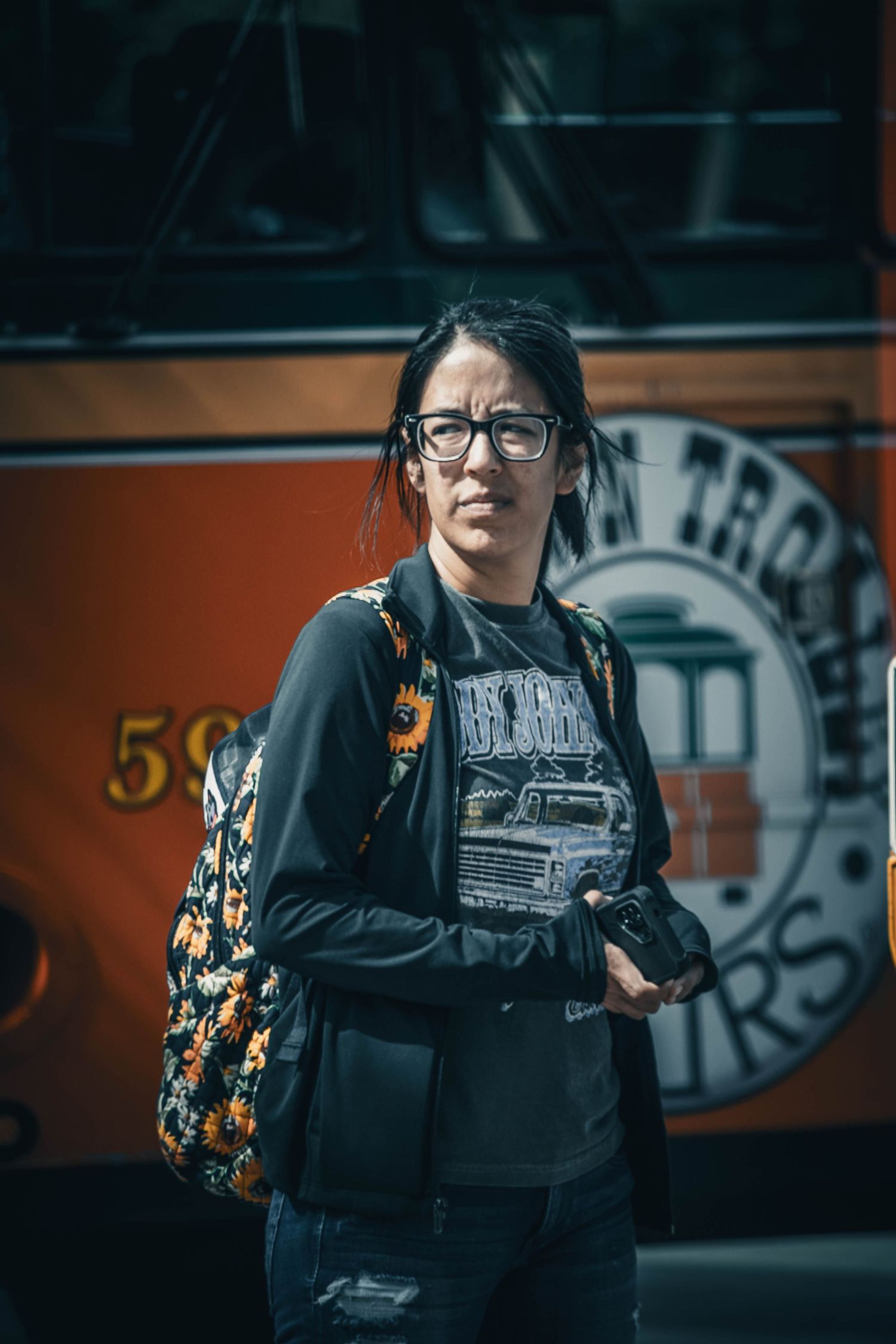 Woman with glasses and floral backpack standing in front of an orange trolley bus on a city street.