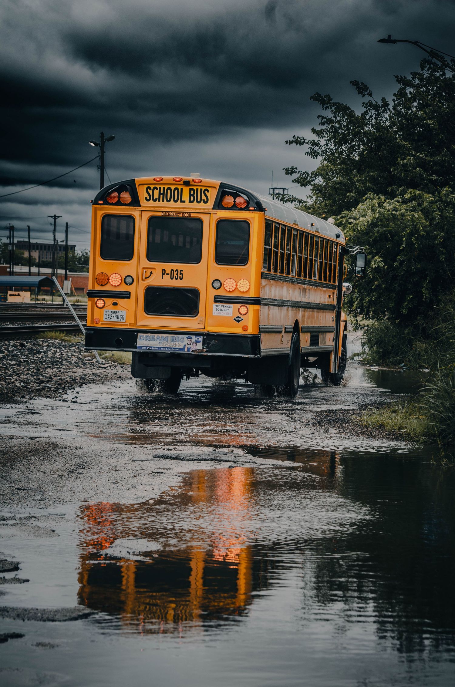 Yellow school bus driving through flooded street under dark stormy clouds with reflection in water.