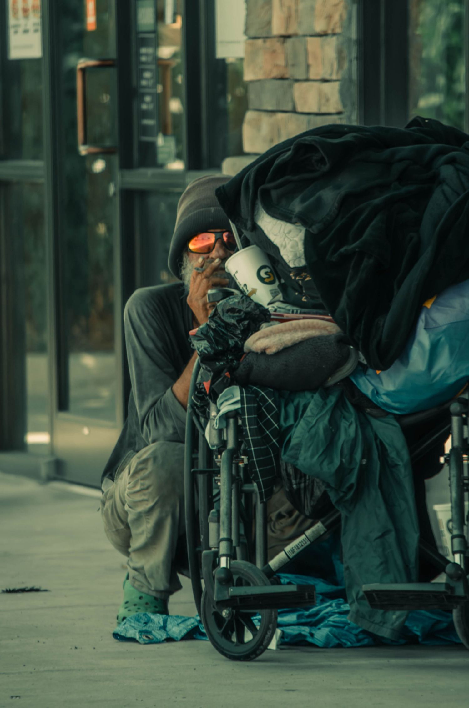 Homeless person crouching beside wheelchair piled with belongings on city sidewalk.