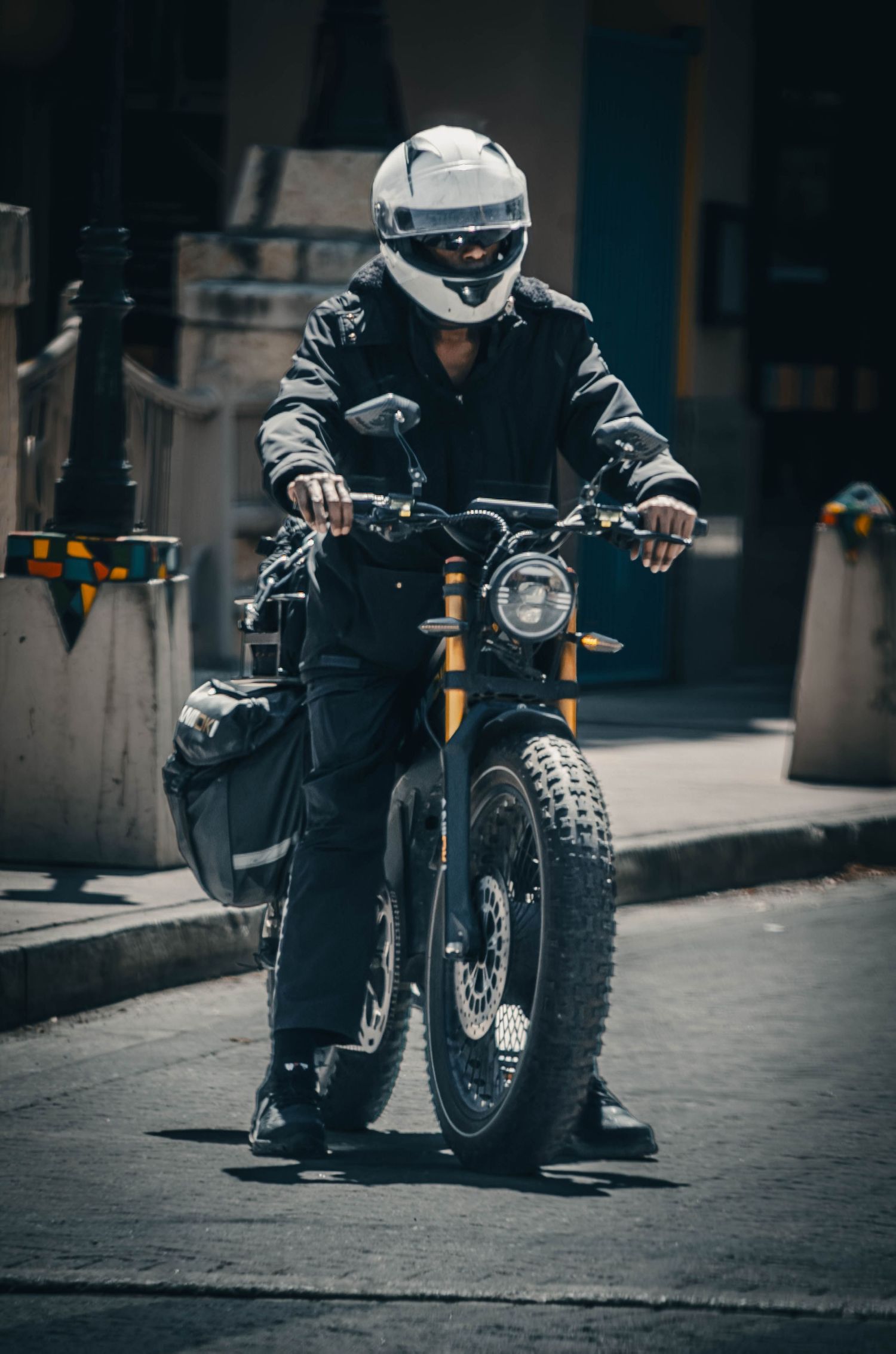 Motorcyclist in full black gear and white helmet riding a scrambler motorcycle on a city street.