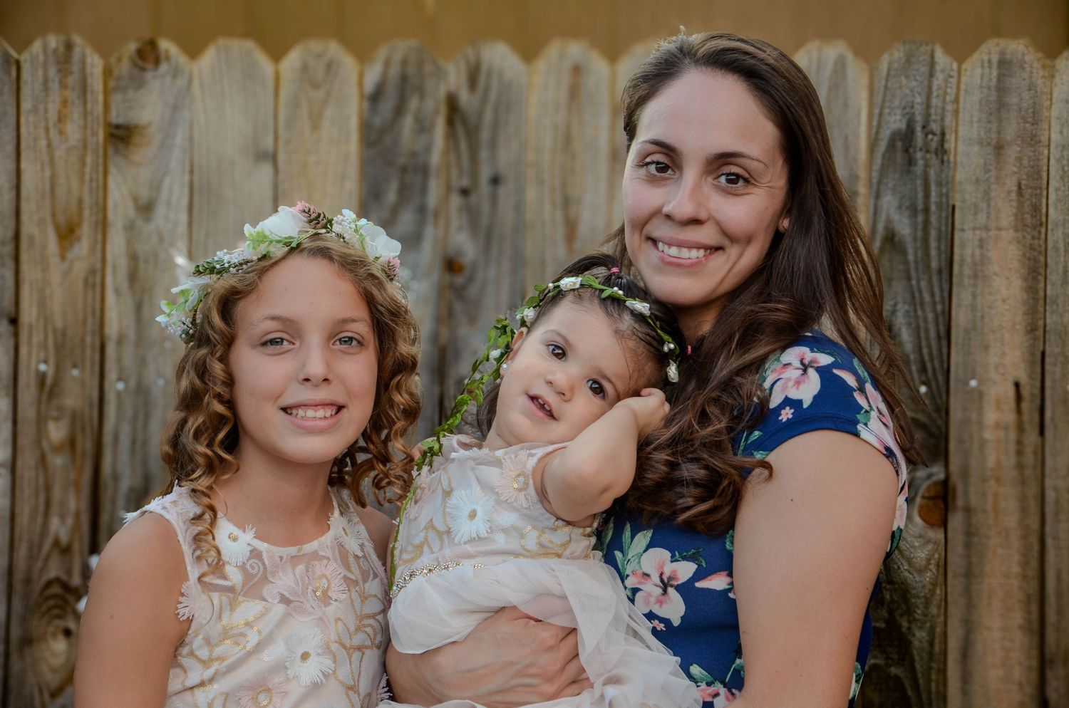 A woman holding a baby and posing with a young girl wearing floral crowns at an outdoor event.