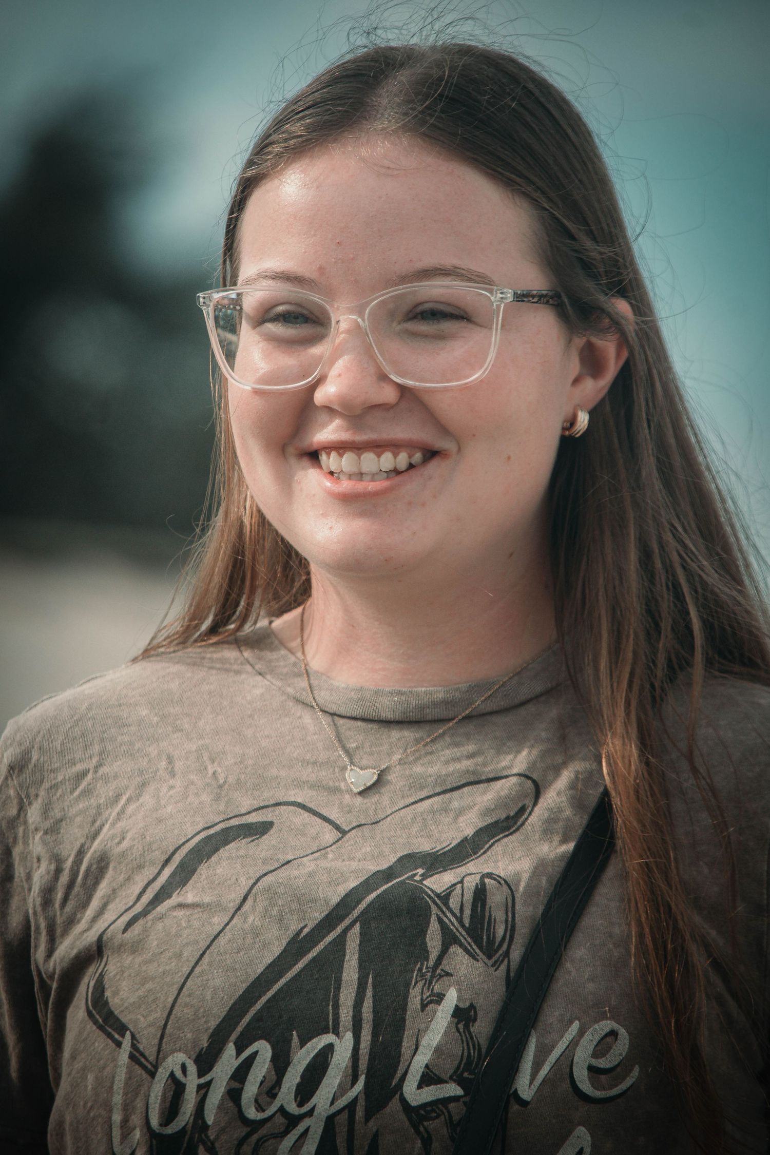 Smiling young woman with glasses and long brown hair wearing a gray graphic tee outdoors.