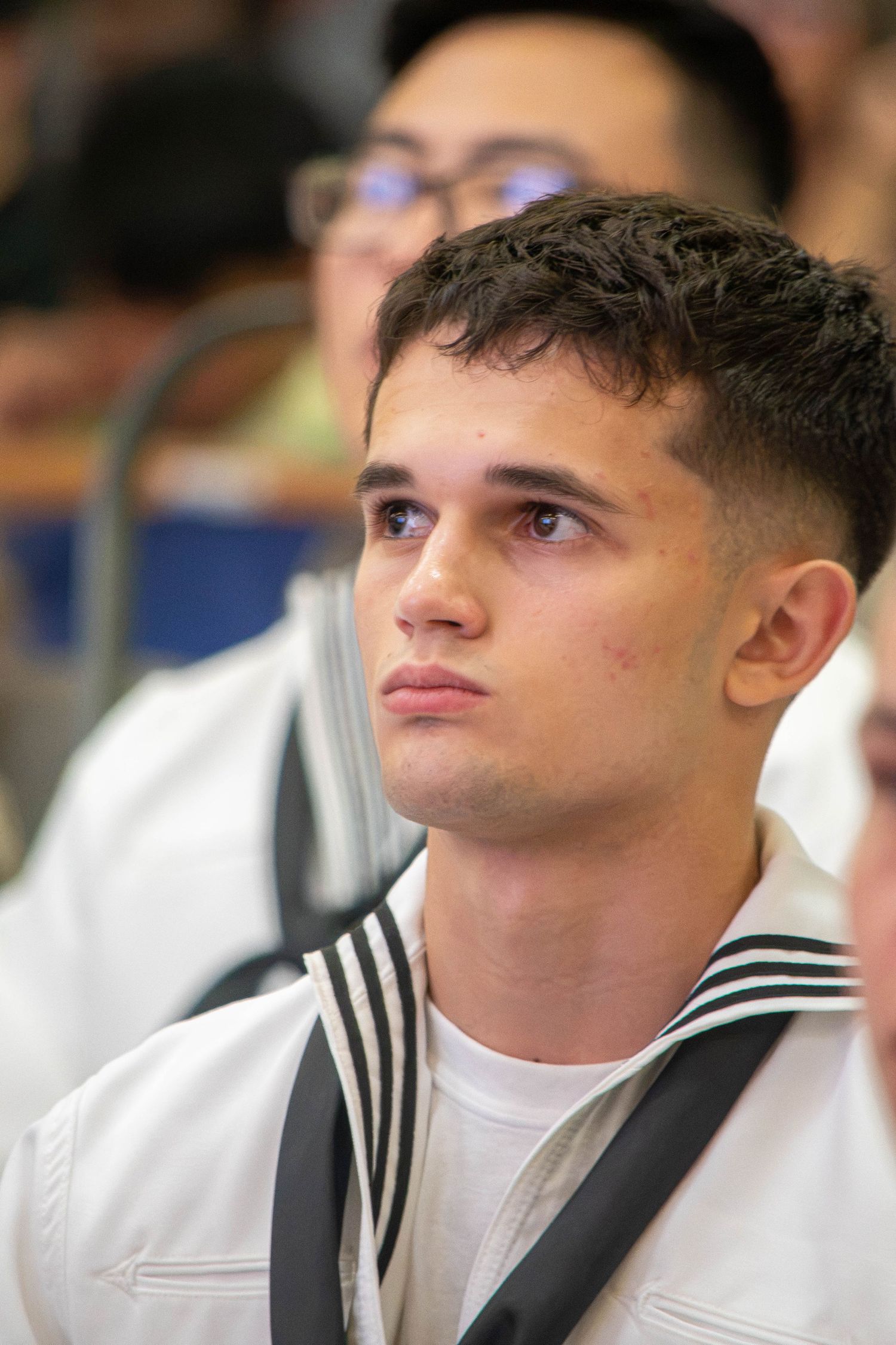 Young male Navy sailor in white uniform gazes upward during an indoor military graduation ceremony.