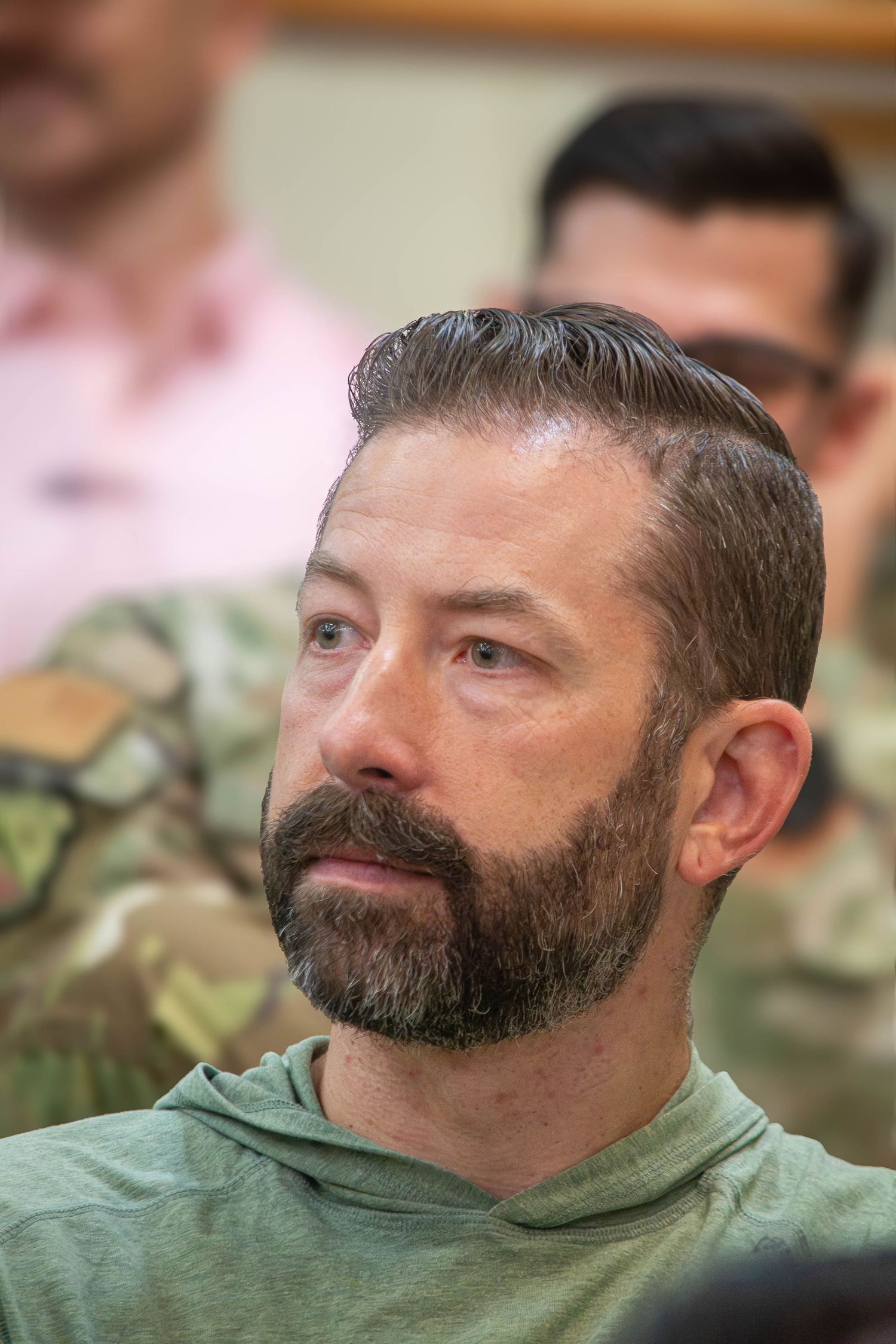 Bearded middle-aged man in green hoodie sitting among military personnel in camouflage uniforms.