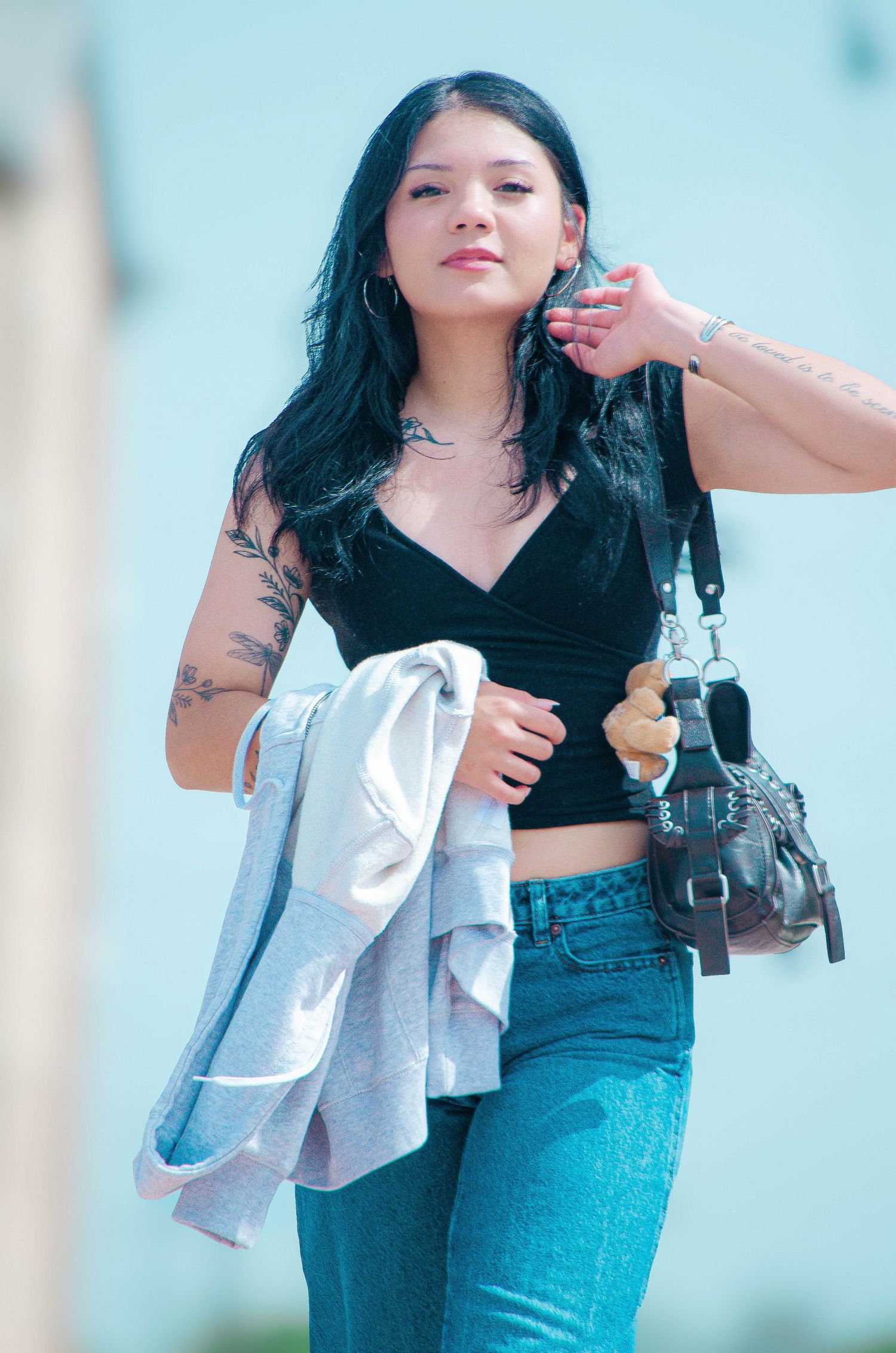 Young woman with tattoos in black crop top and jeans carrying jacket and bag outdoors.