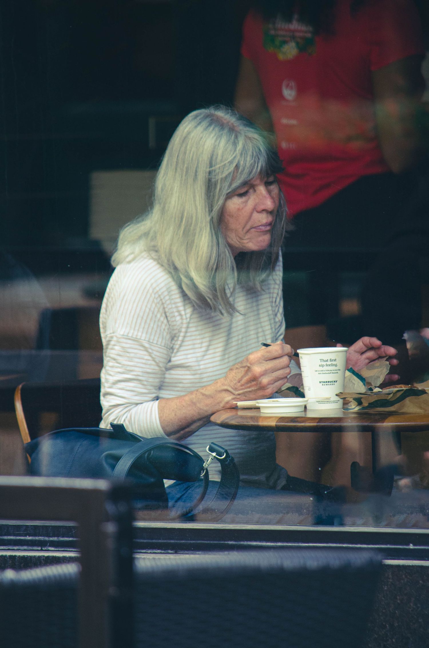 Older woman with long gray hair sitting alone at a cafe table with a coffee cup.