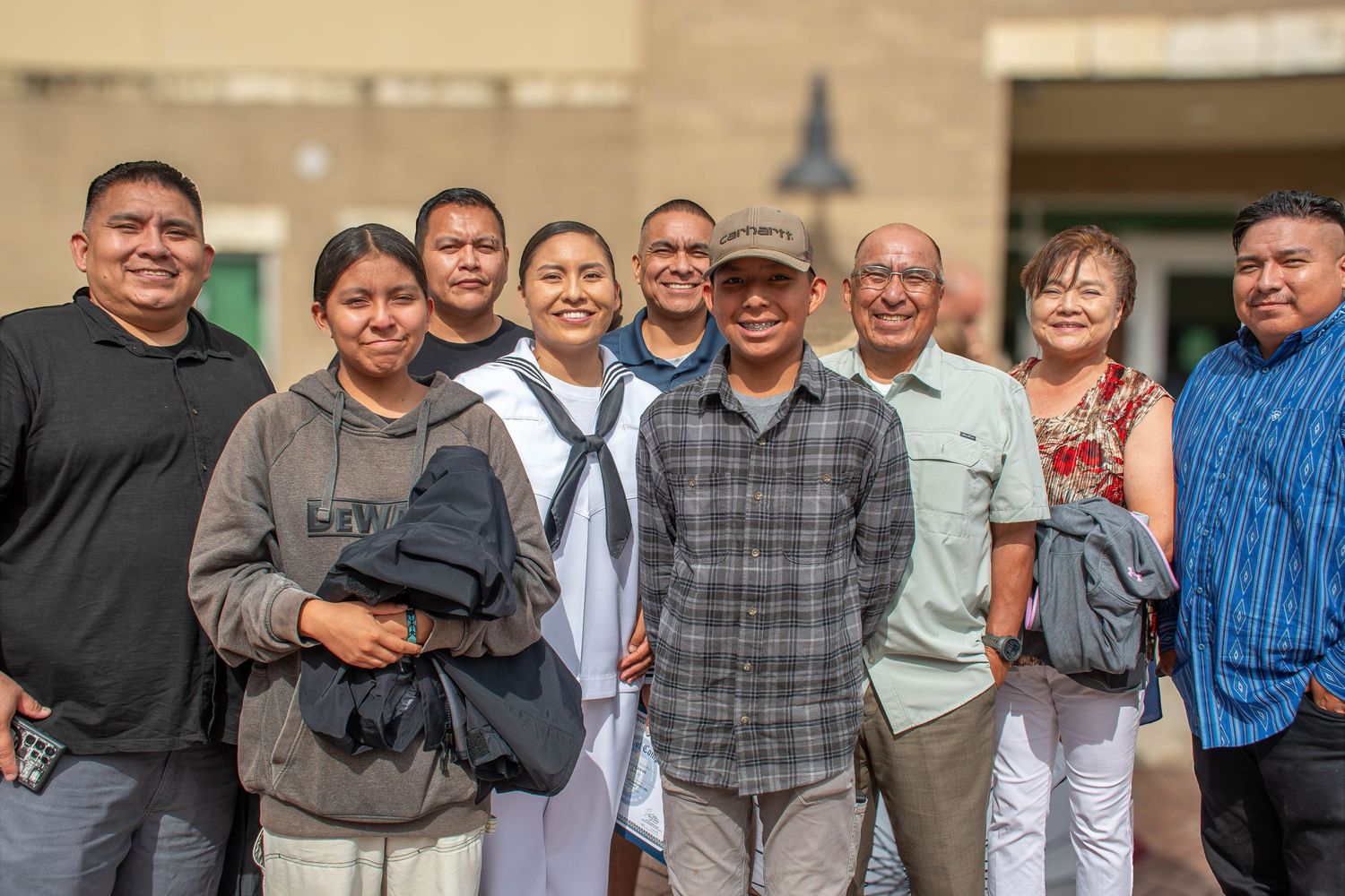 Navy sailor in dress whites poses with large family group outside military building.