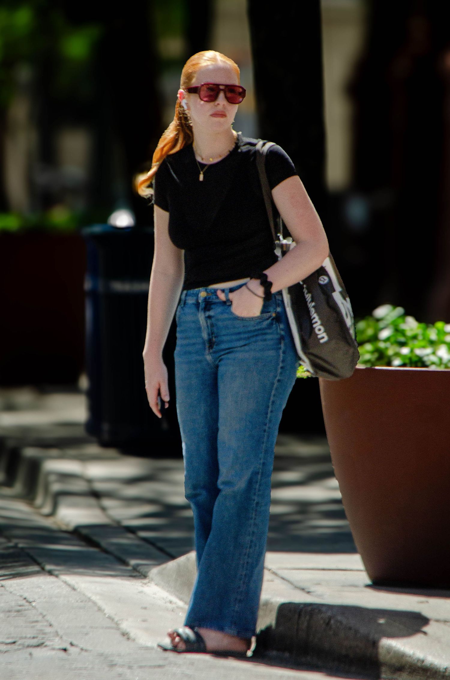 A young redhead woman in a black t-shirt and wide-leg jeans walking on a sunny urban sidewalk.