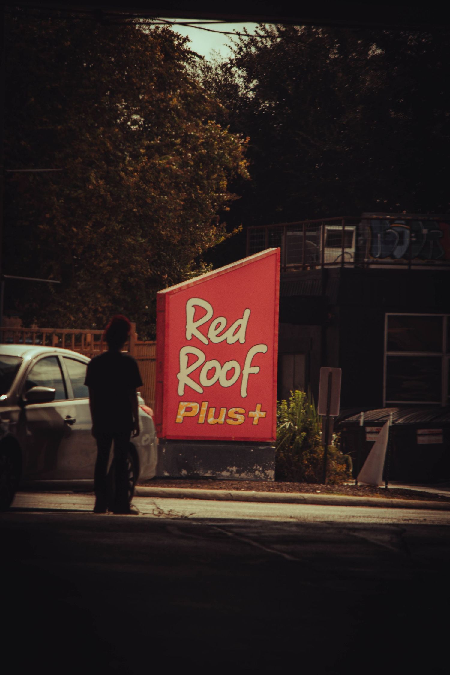 Person standing near a Red Roof Plus+ hotel sign in a parking lot surrounded by trees at dusk.