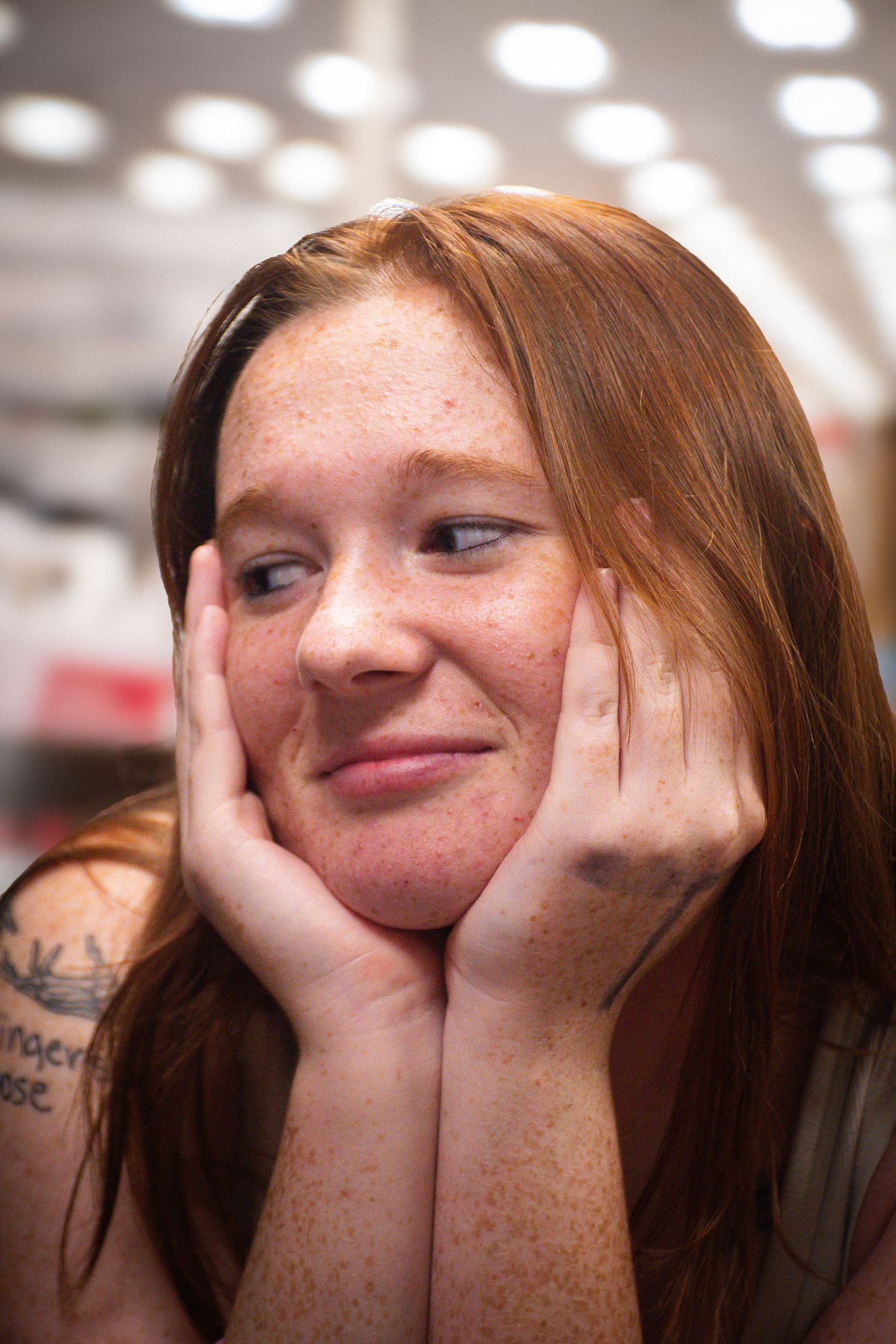 Smiling redhead woman with freckles resting her face in her hands, wearing a tank top with a visible tattoo.
