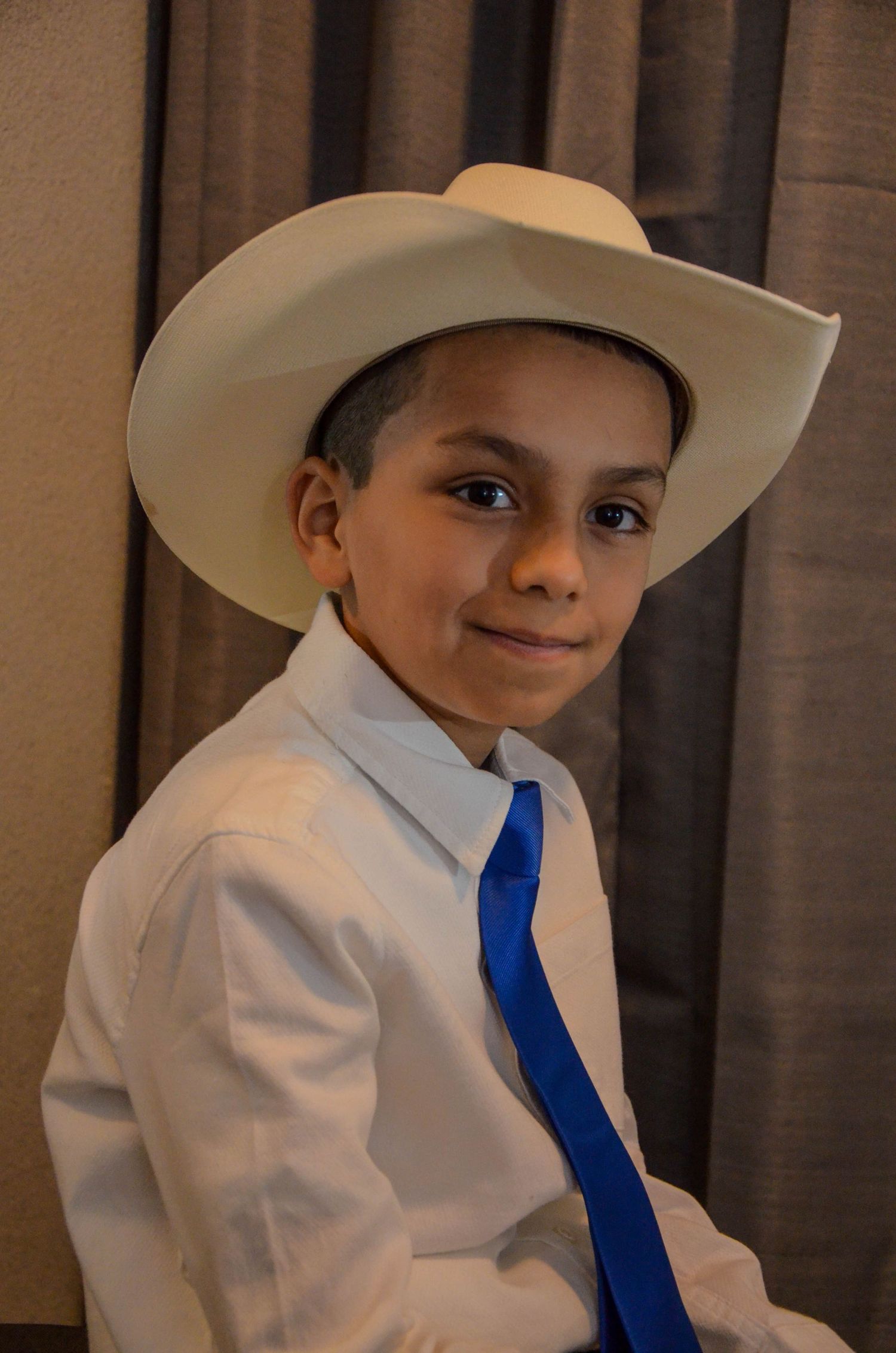 Young boy wearing a white cowboy hat and blue tie with white dress shirt.