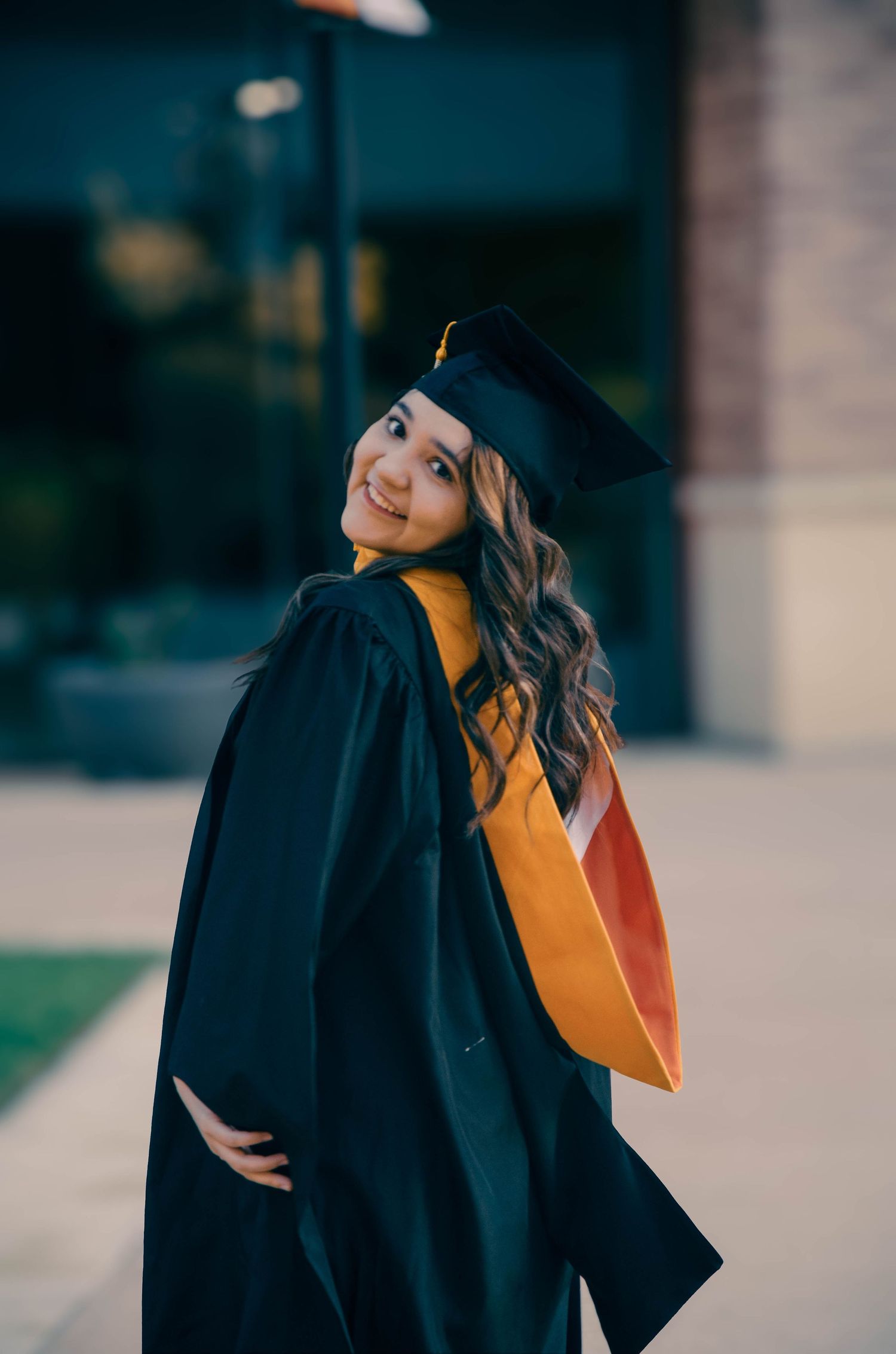 Smiling female graduate in black cap and gown with gold sash posing outdoors.