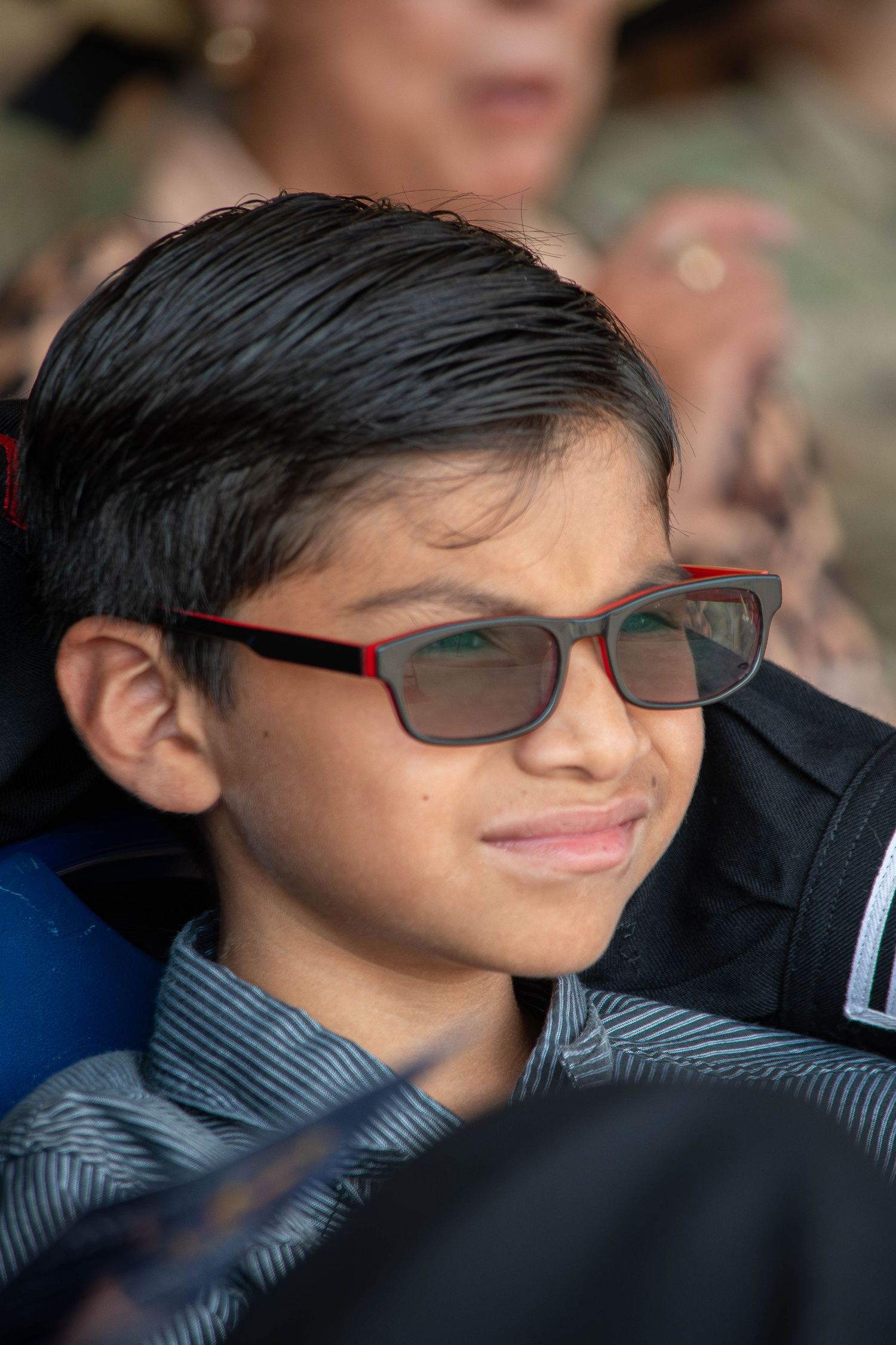 A young boy wearing red-framed glasses smiles softly at an outdoor military event.