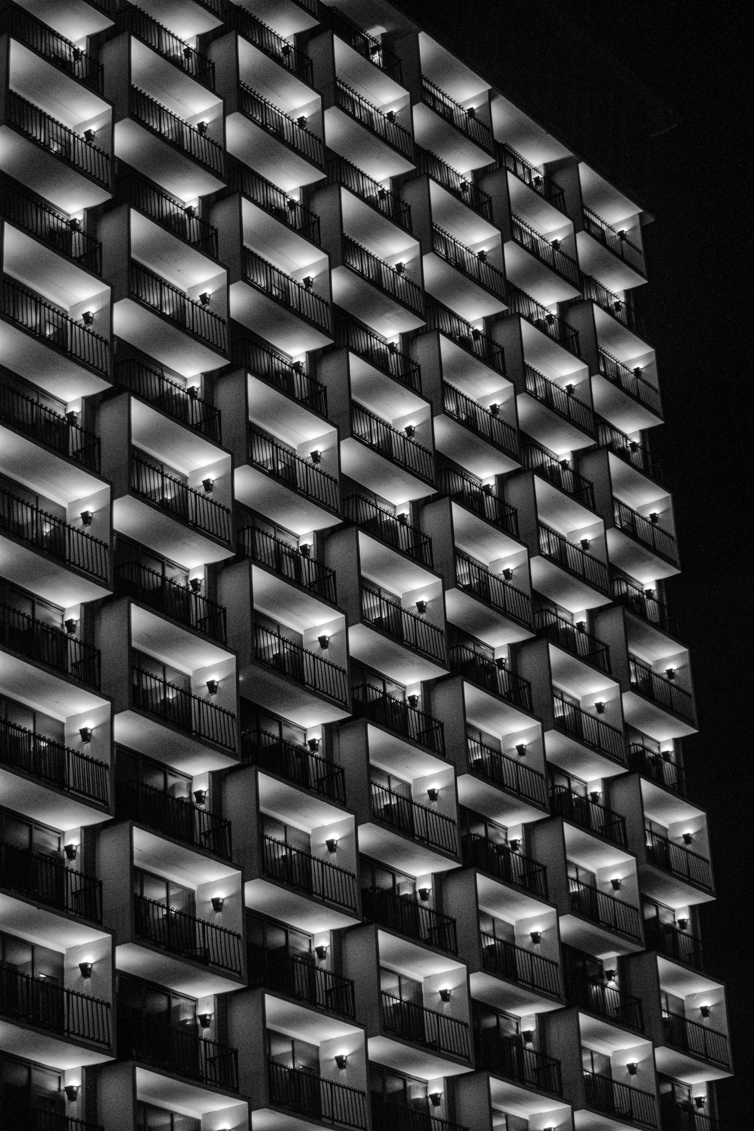 Black and white abstract pattern of illuminated hotel balconies creating a geometric diamond grid at night.