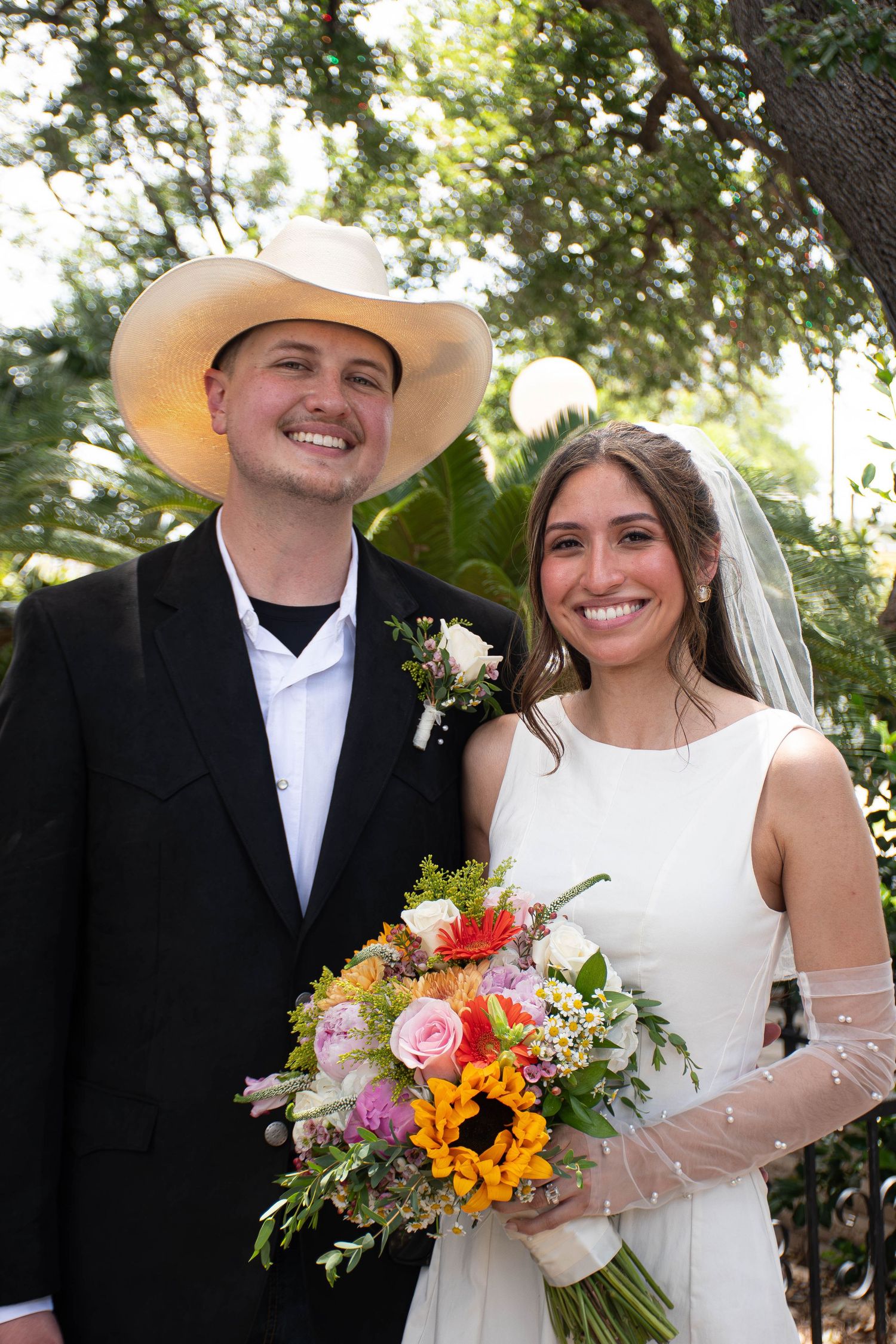 Smiling bride and groom in cowboy hat holding colorful wedding bouquet outdoors.