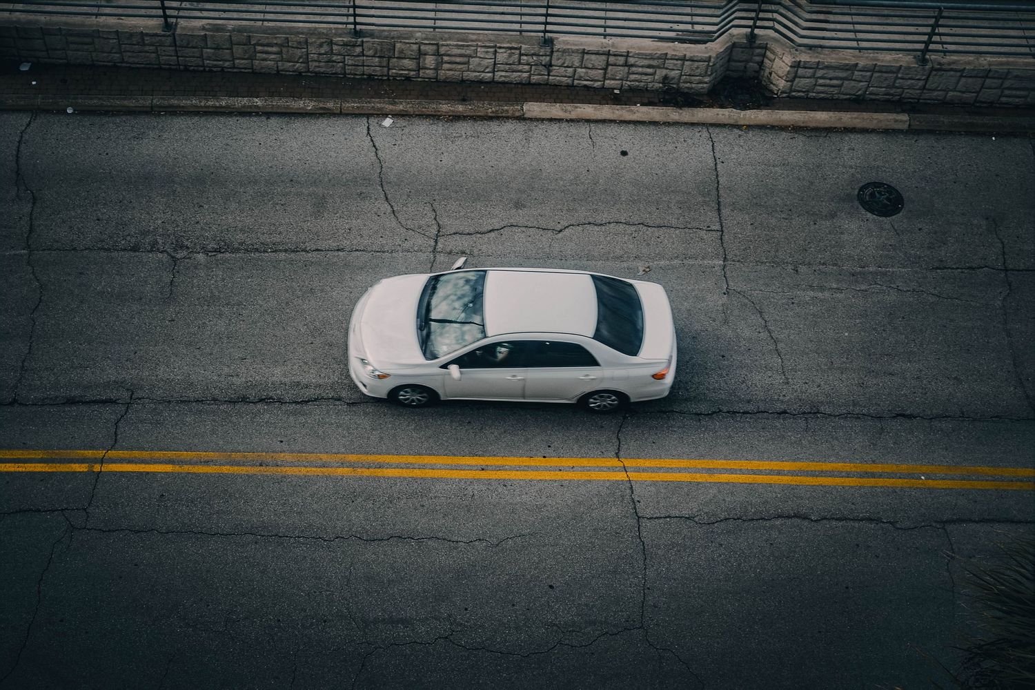 Aerial view of a white sedan driving on a cracked asphalt road with yellow center lines.