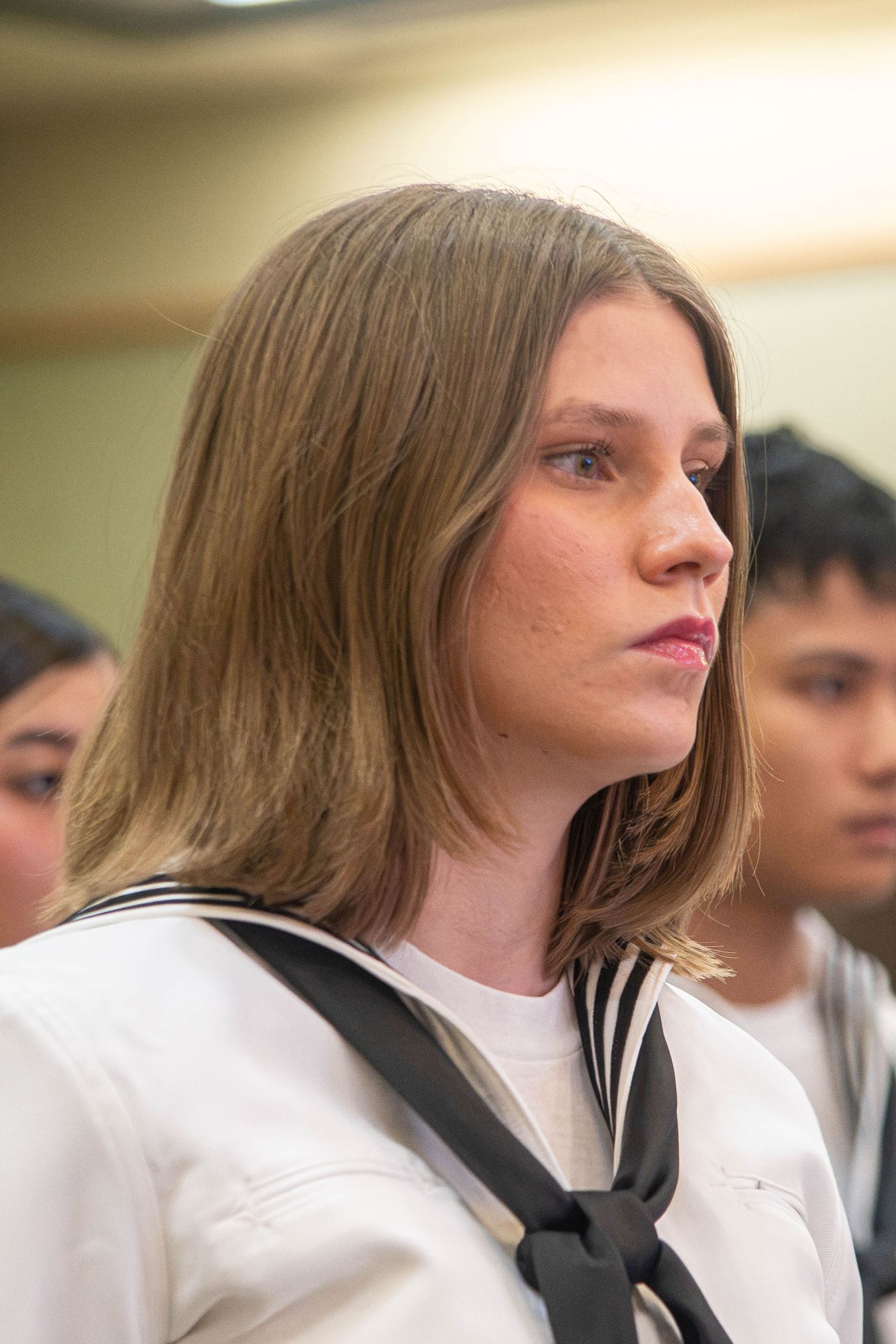Young female Navy sailor with long blonde hair in white uniform attends military graduation ceremony indoors.
