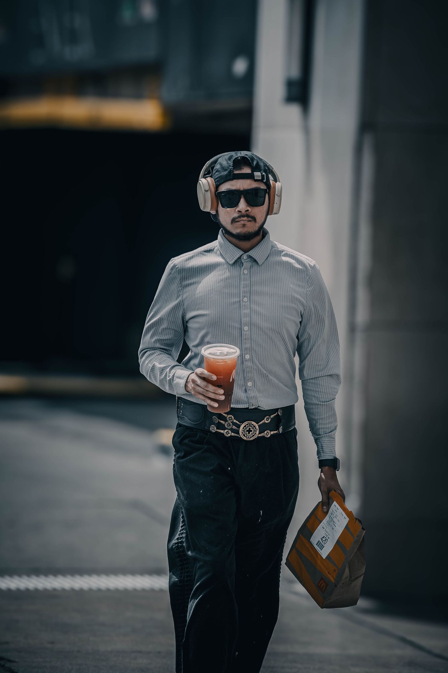 Stylish man walking outdoors wearing headphones, sunglasses, holding a drink and carrying a box.