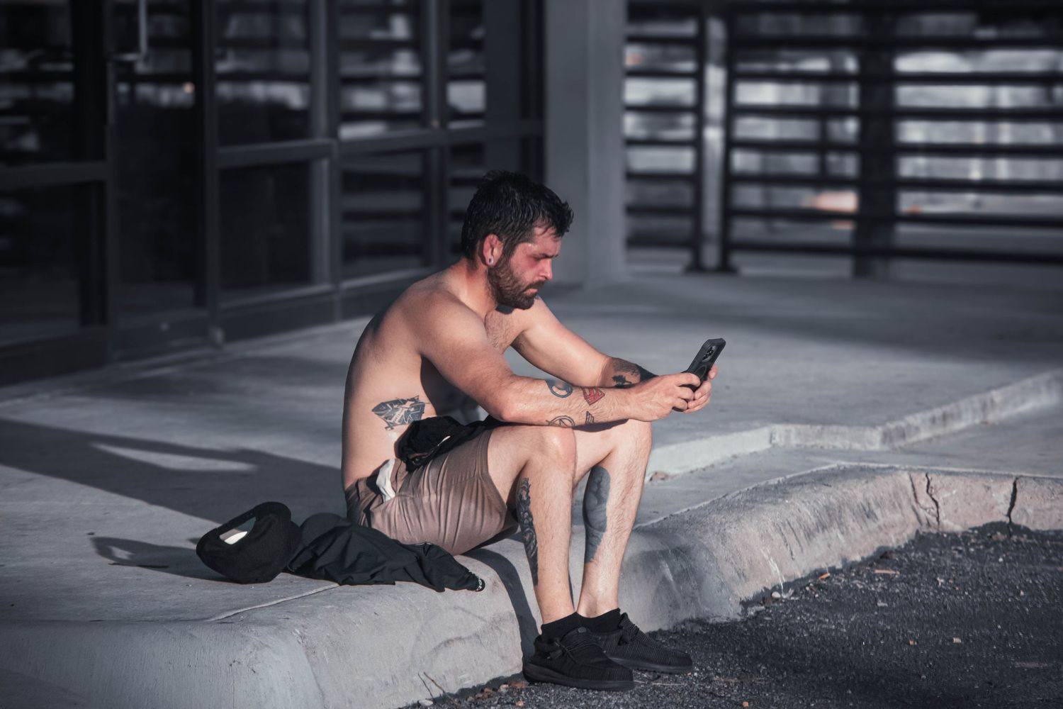 Shirtless bearded man sitting on a curb outdoors at night, looking at his smartphone.