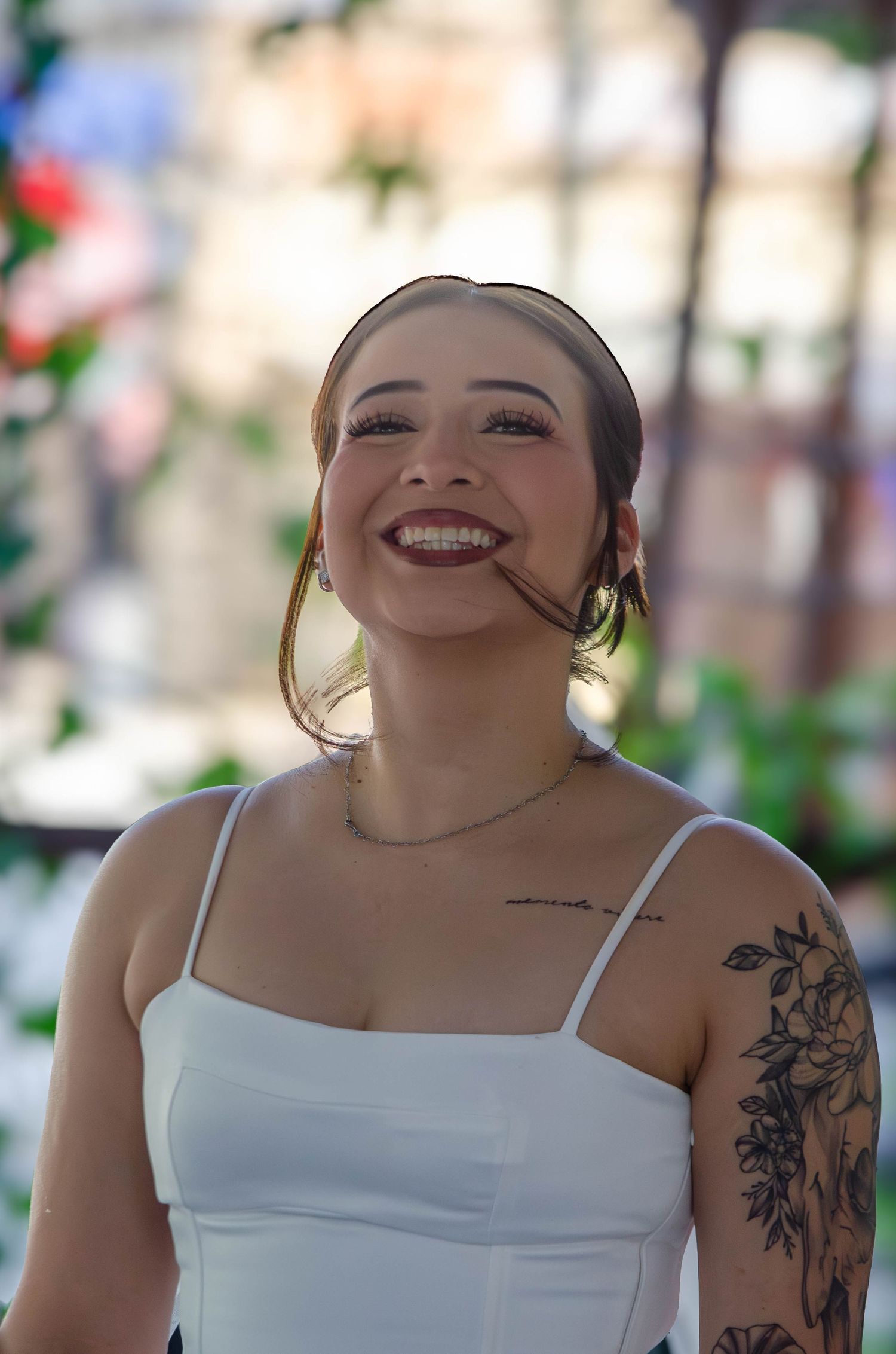 Smiling young woman with floral arm tattoo and dark lipstick wears white top outdoors at a celebration.