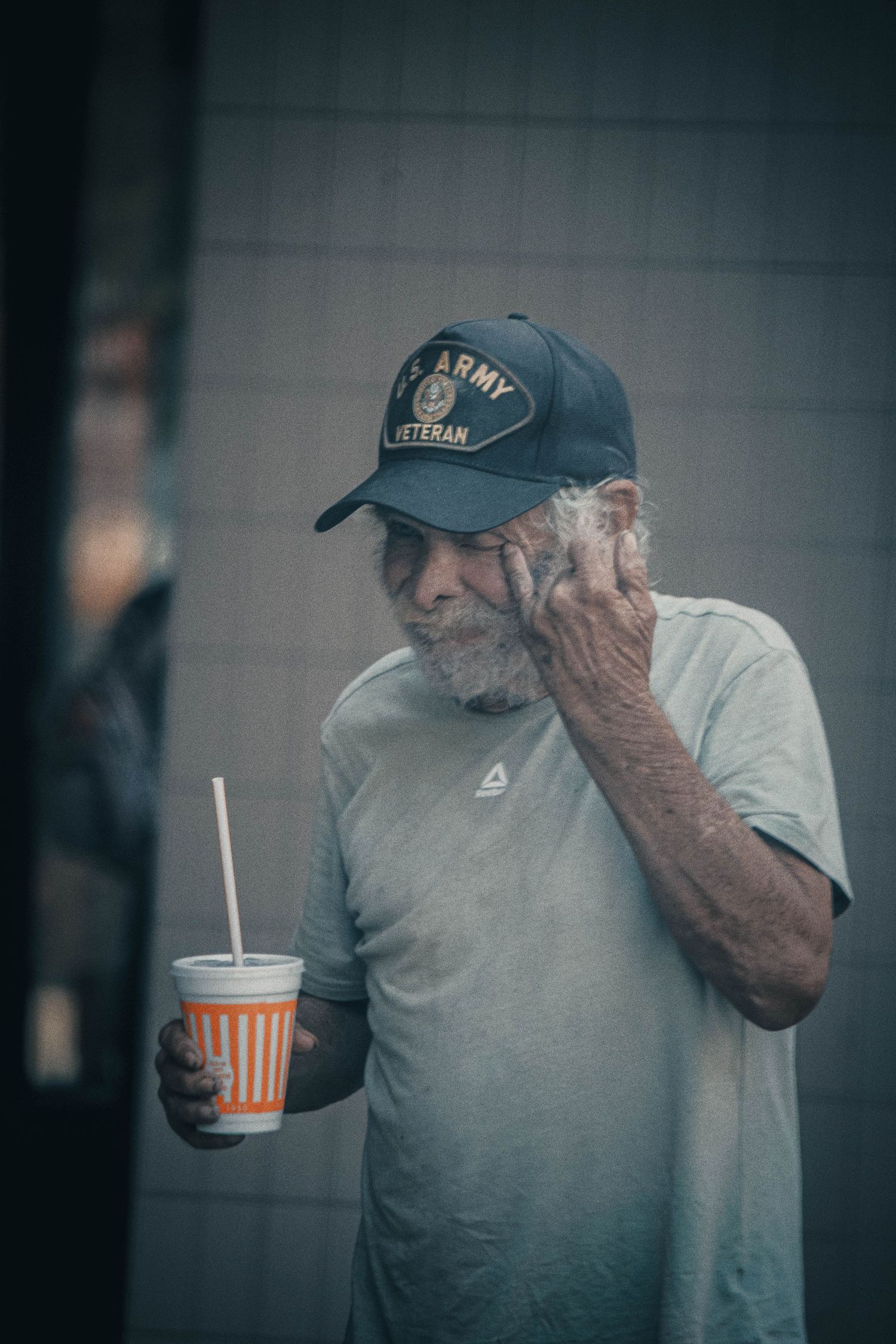 Elderly US Army veteran wearing a military cap holds a striped cup with a straw outdoors.