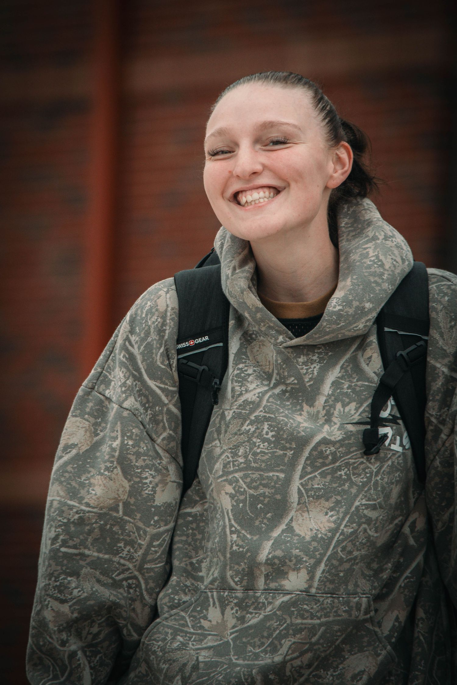 Smiling young woman wearing a patterned oversized hoodie and black backpack standing outdoors near a brick wall.