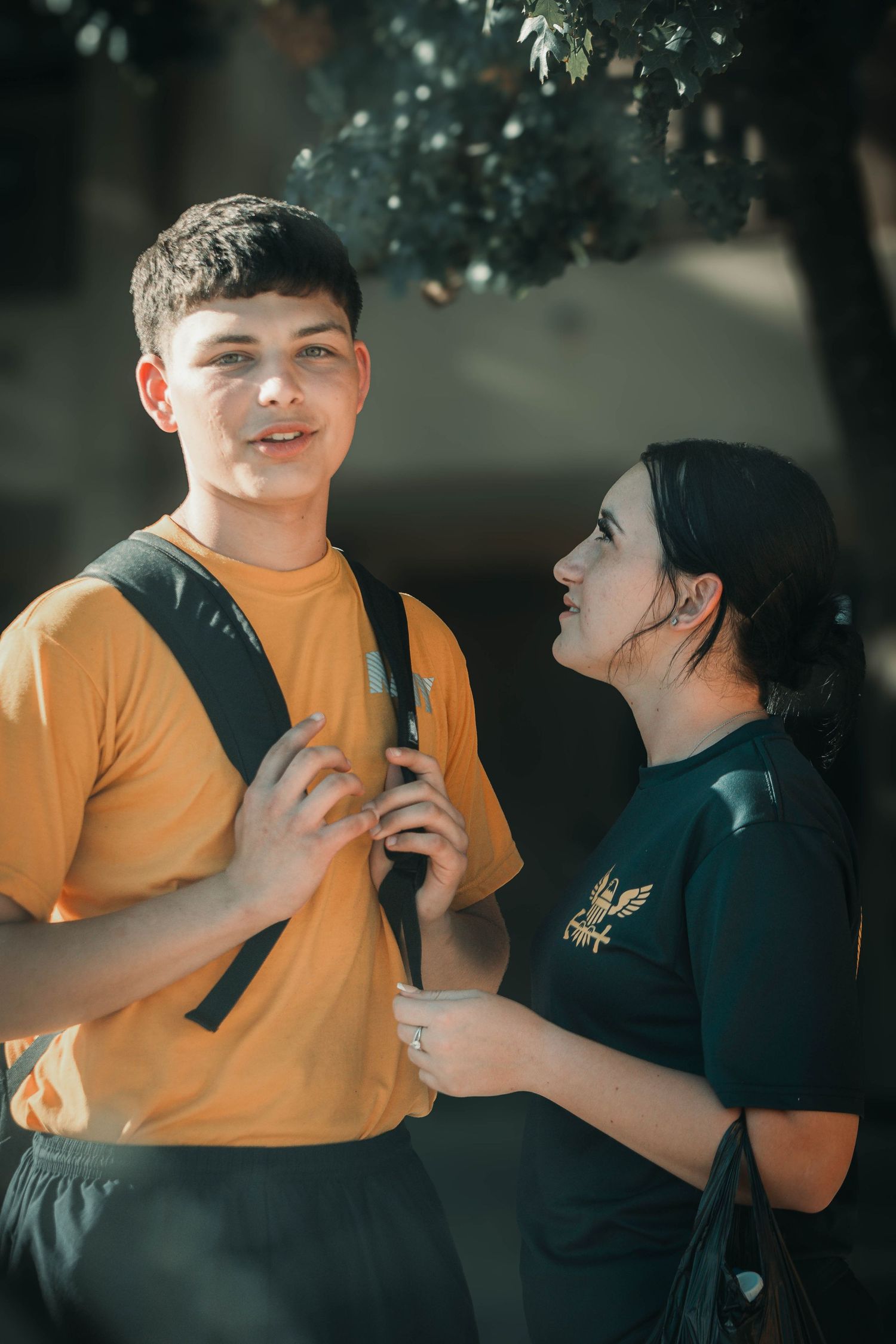 Two young students share a moment outdoors, boy in yellow shirt with backpack smiling at dark-haired girl in black tee.
