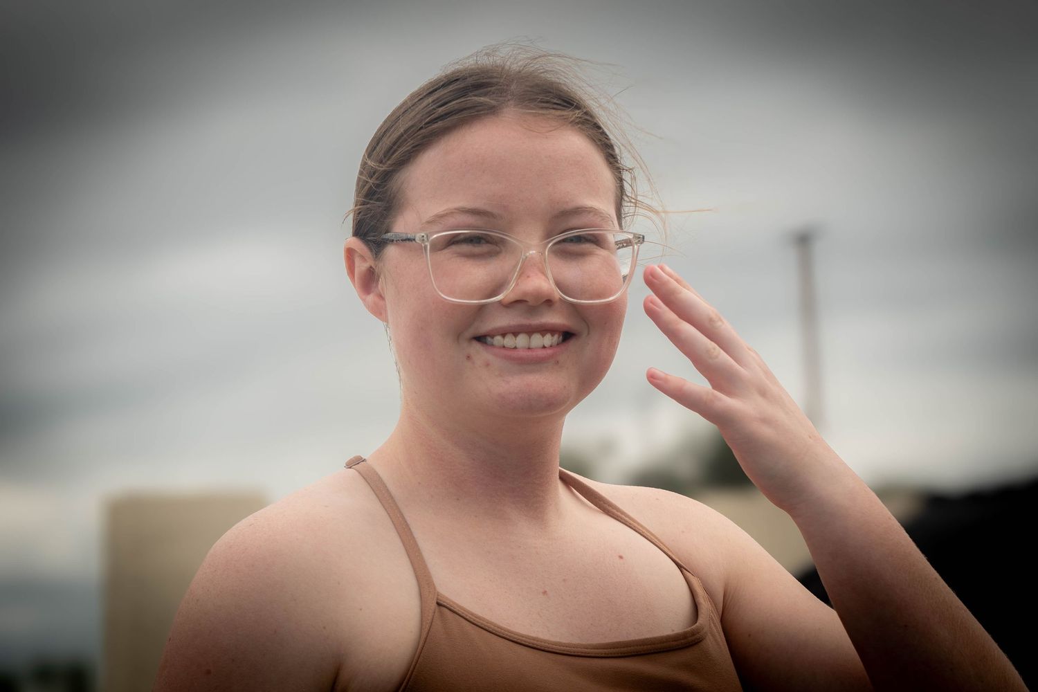Smiling young woman with glasses making a peace sign outdoors on a cloudy day.
