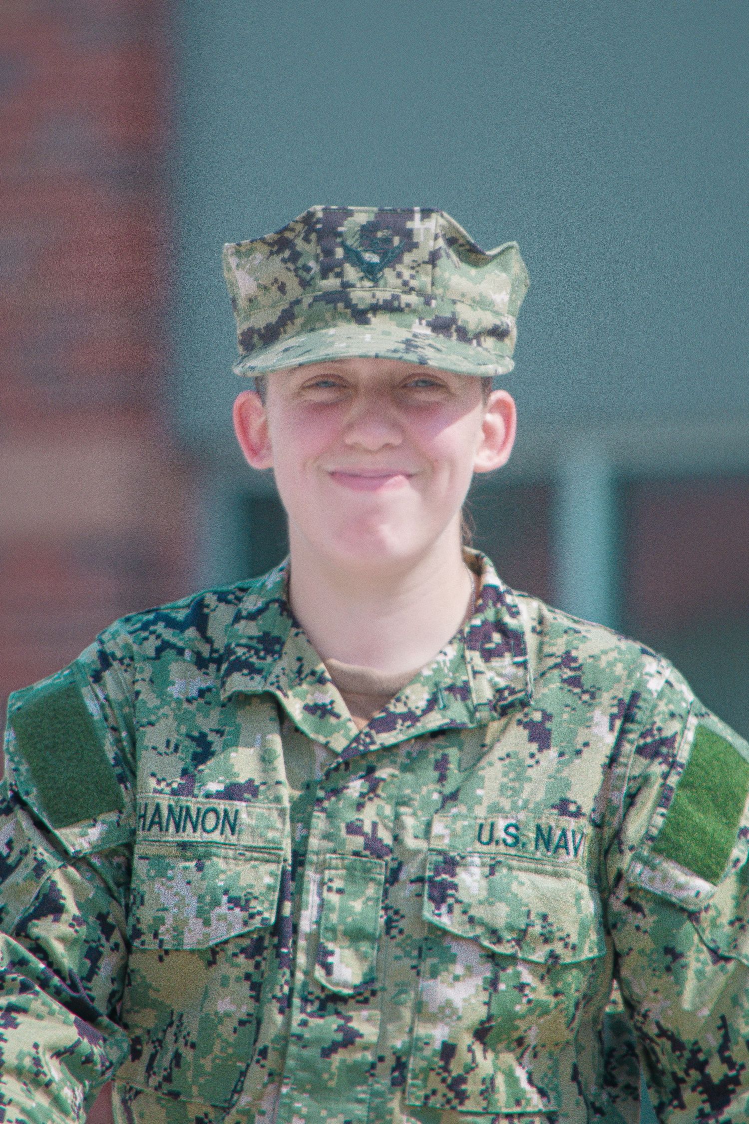 U.S. Navy sailor in digital camouflage uniform and cap smiling outdoors near a building.