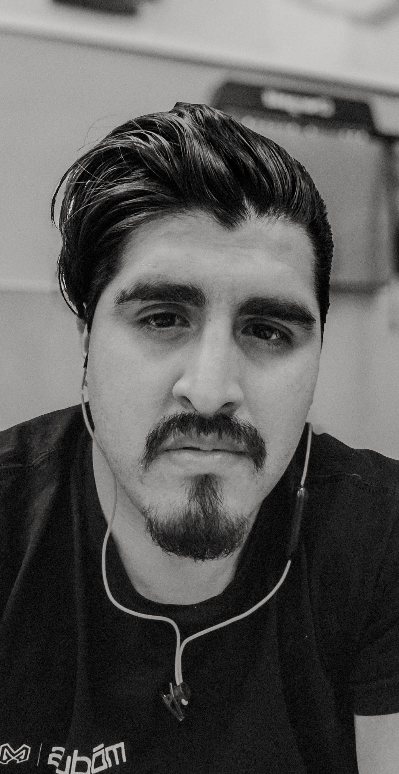 Black and white selfie of a young man with slicked-back hair, goatee, and earbuds wearing a black t-shirt.