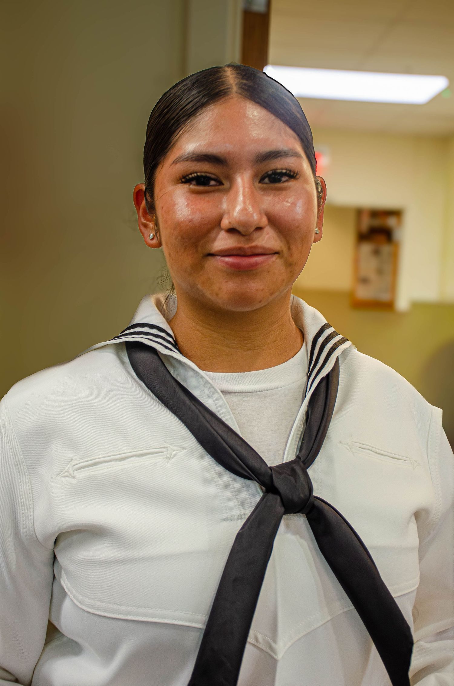 Young US Navy sailor in white dress uniform with black neckerchief smiling indoors.