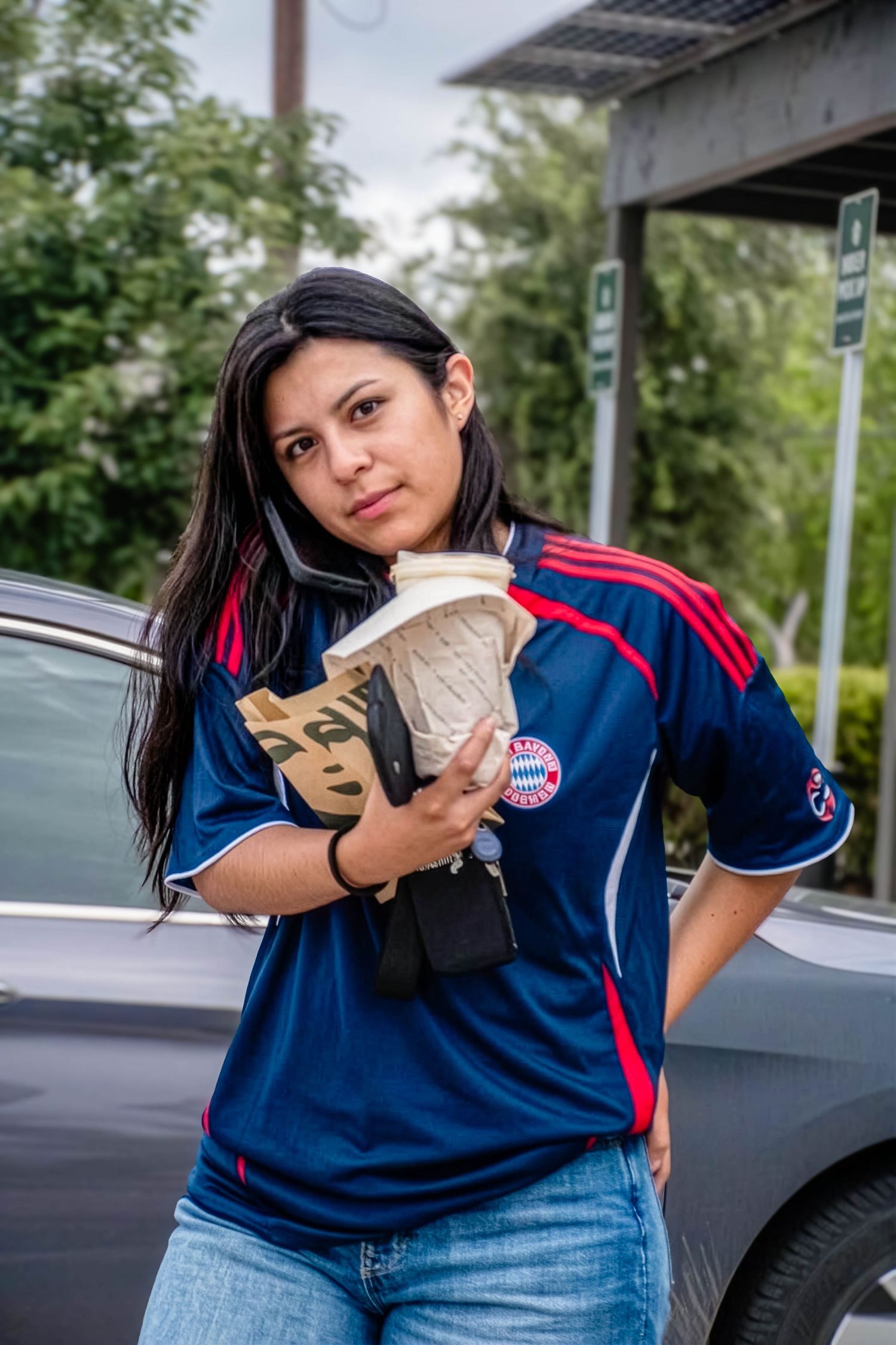 Young woman in Bayern Munich jersey holding coffee cup standing near car outdoors.