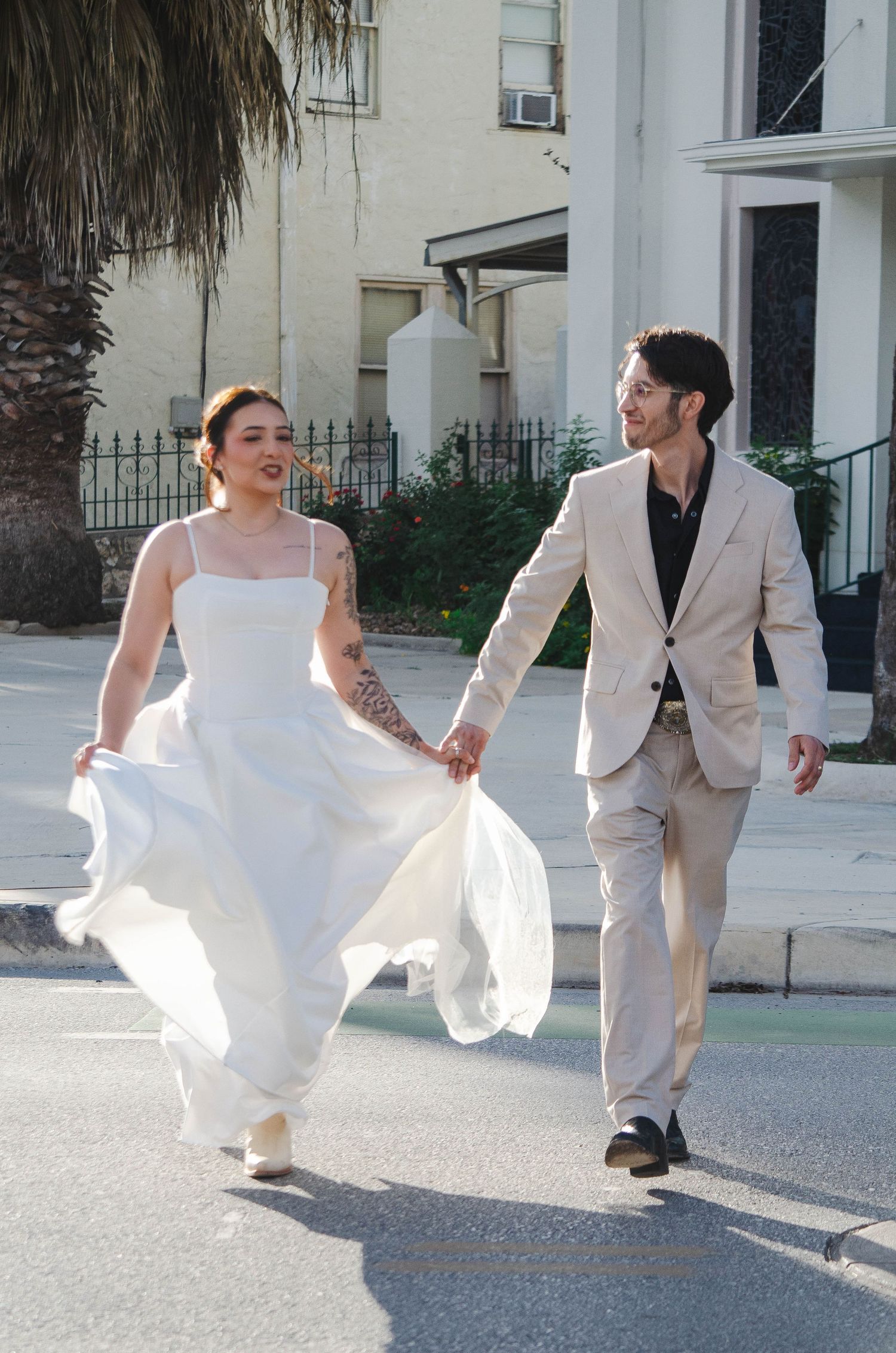 Bride in flowing white gown and groom in beige suit walk hand in hand down a sunny street on wedding day.