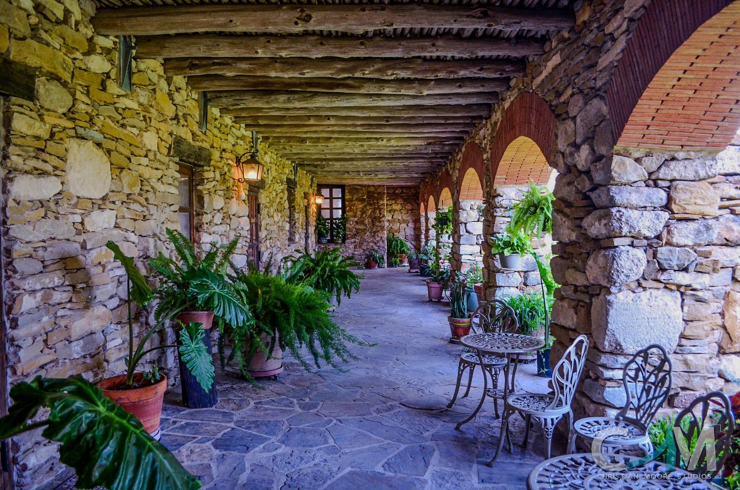 Rustic stone archway corridor with lush ferns, potted plants, and wrought iron furniture at a historic venue.