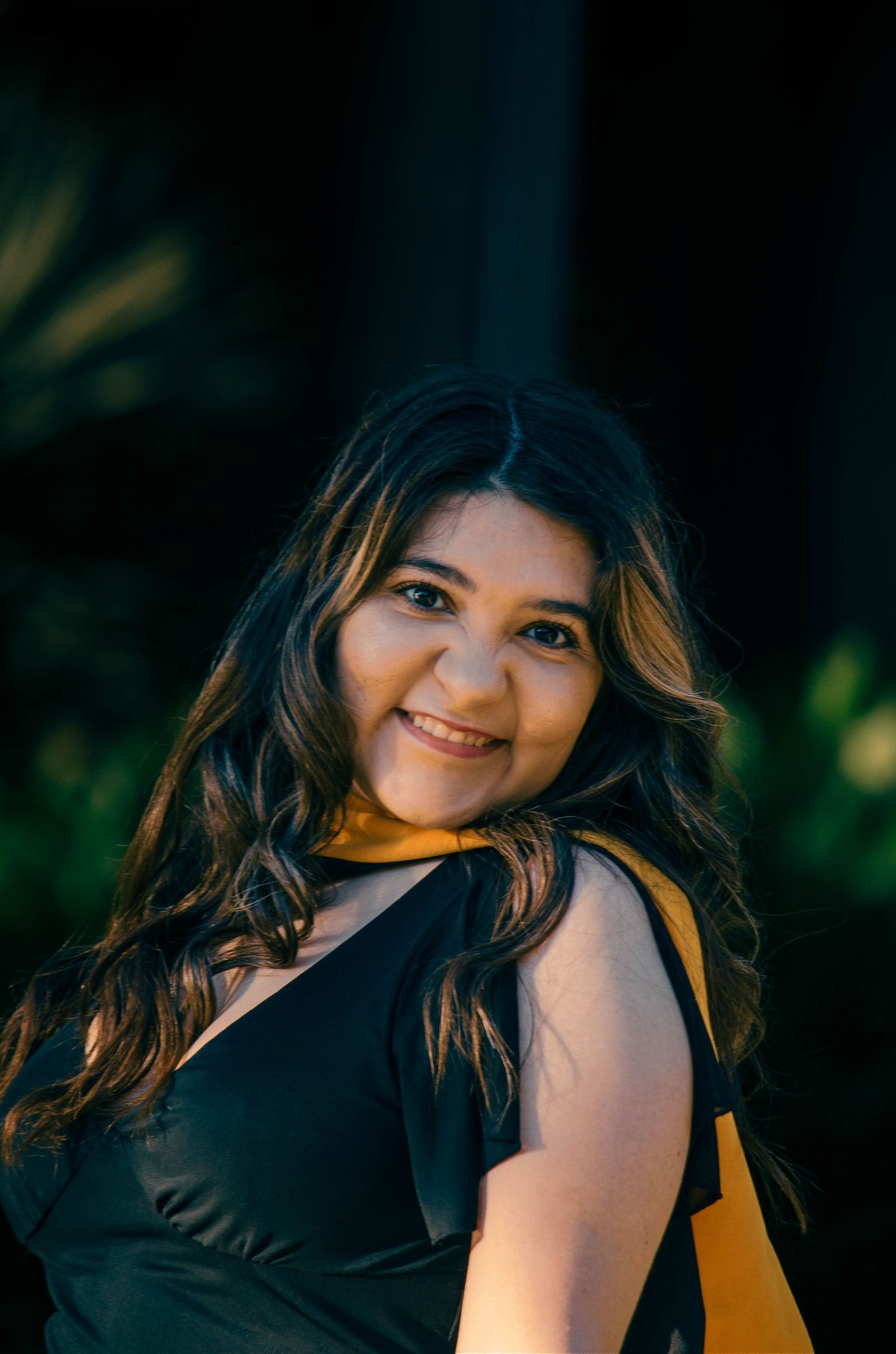 A smiling young woman with long wavy hair wearing a black and yellow dress outdoors at night.