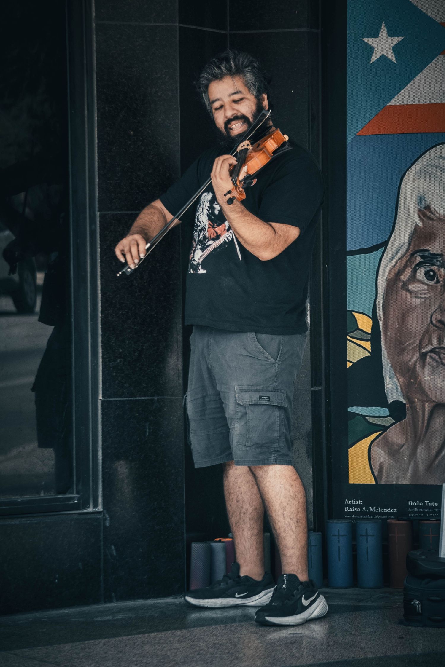 Street musician playing violin outdoors near a colorful mural, wearing black t-shirt and gray shorts.