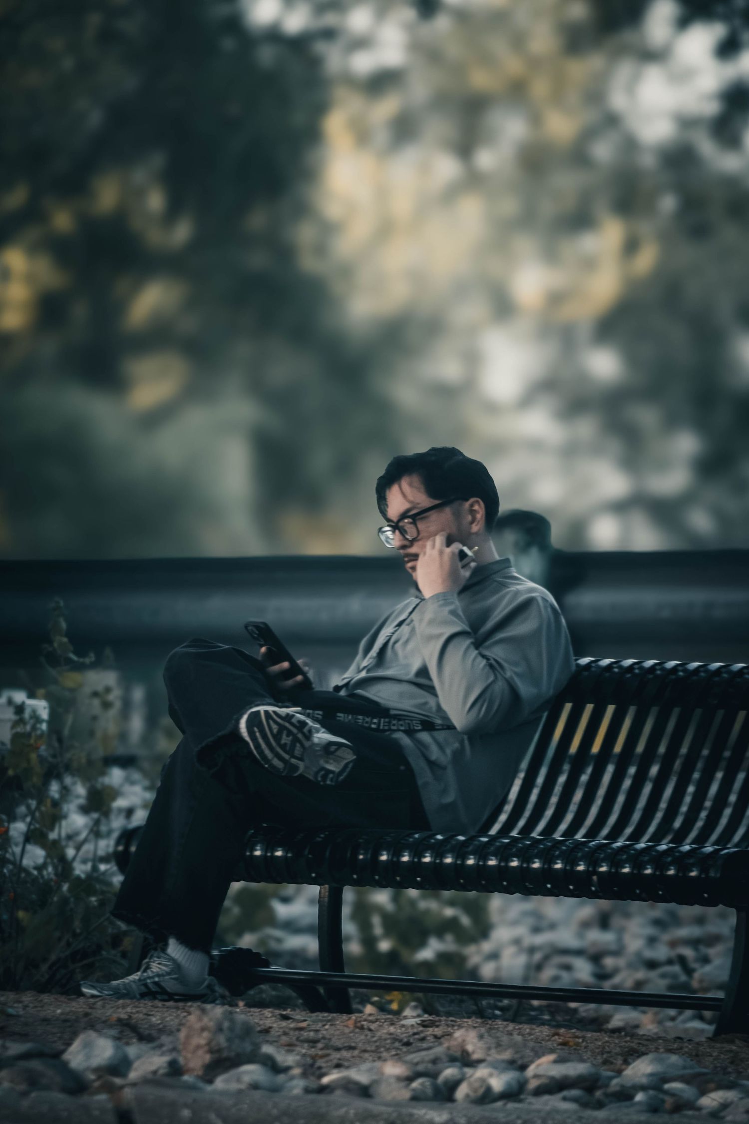 Man with glasses sitting on a park bench reading a tablet, surrounded by trees in a moody outdoor setting.