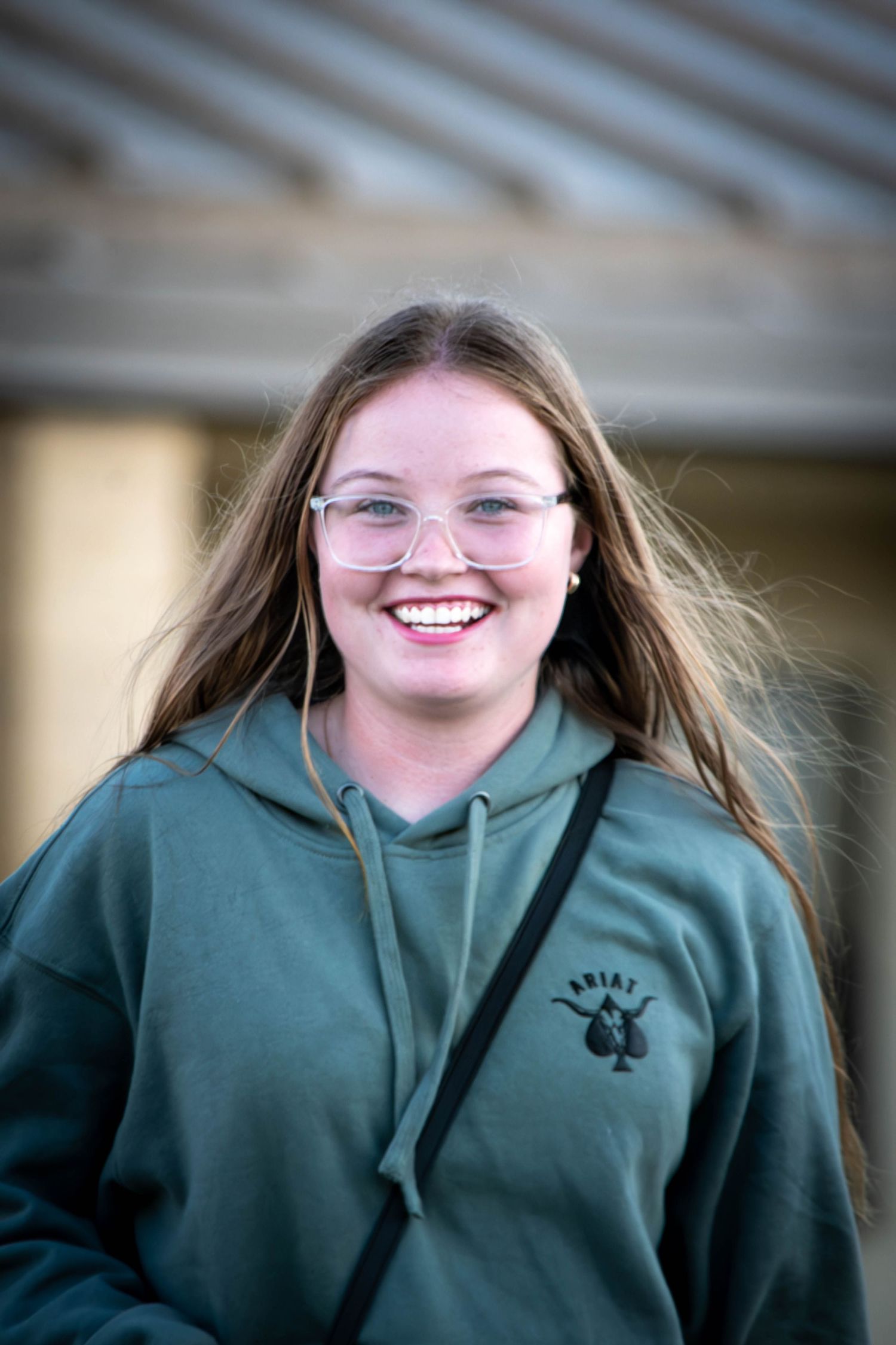 Smiling young woman with long hair and clear glasses wearing a green Ariat hoodie outdoors.