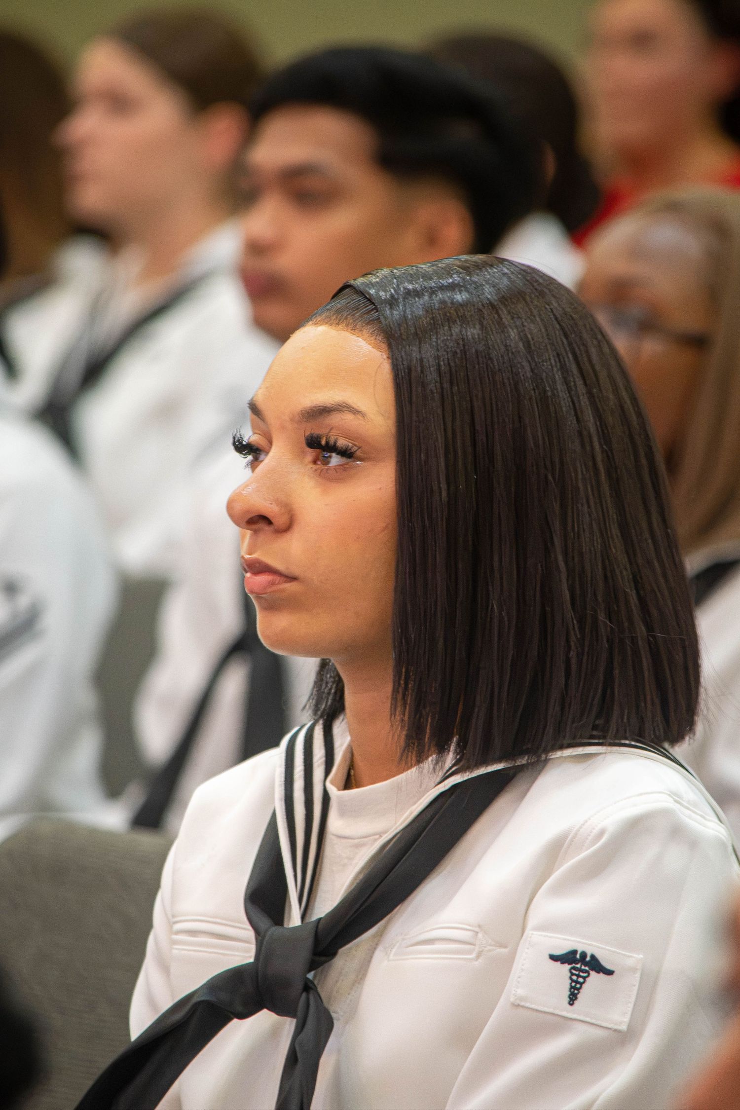 Female Navy sailor with bob haircut in white uniform and medical insignia attends military graduation ceremony.