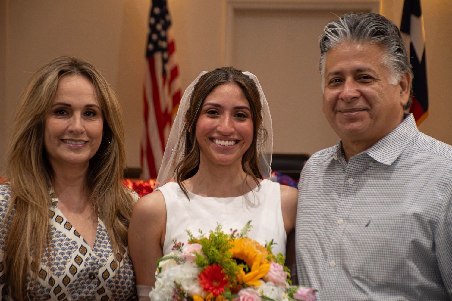 Bride with colorful bouquet posing with parents in front of American flag indoors.